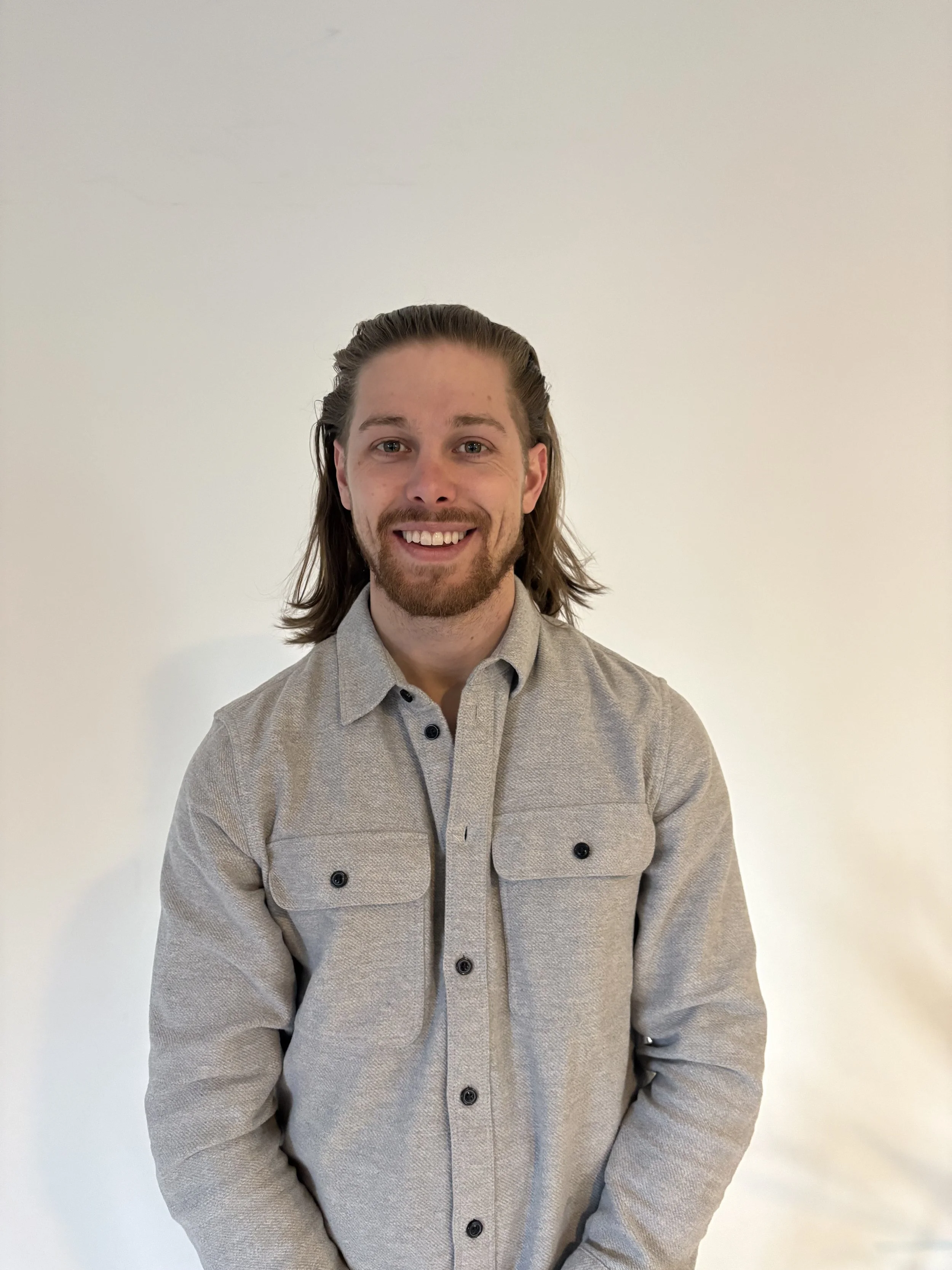 A man with shoulder-length brown hair, a beard, and a mustache, smiling, wearing a light gray button-up shirt with two chest pockets, standing against a plain white wall.