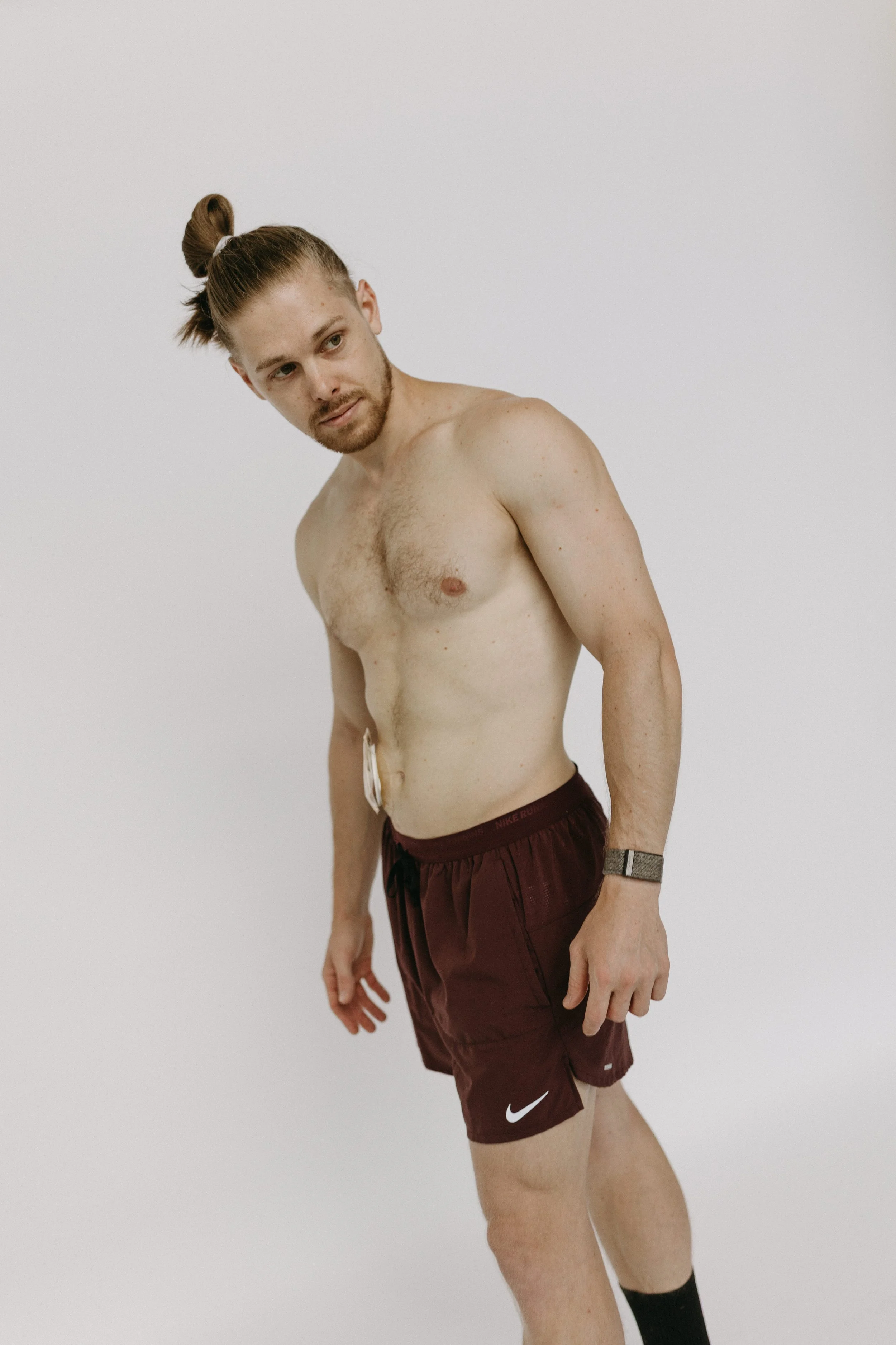A shirtless man with long brown hair tied in a bun, wearing maroon Nike running shorts and a watch, standing against a plain white background.