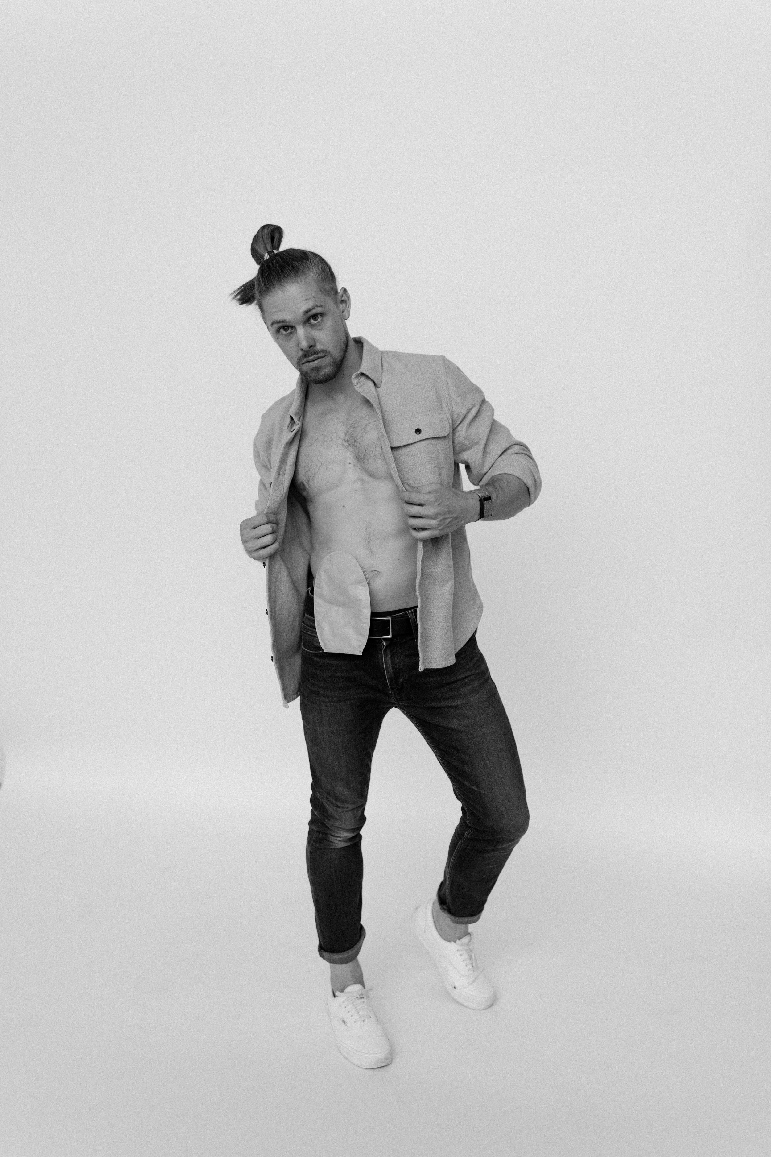 Black and white photo of a man standing with his shirt open, exposing his chest and abdomen, wearing jeans and sneakers, with one hand adjusting his shirt and the other holding open his shirt.