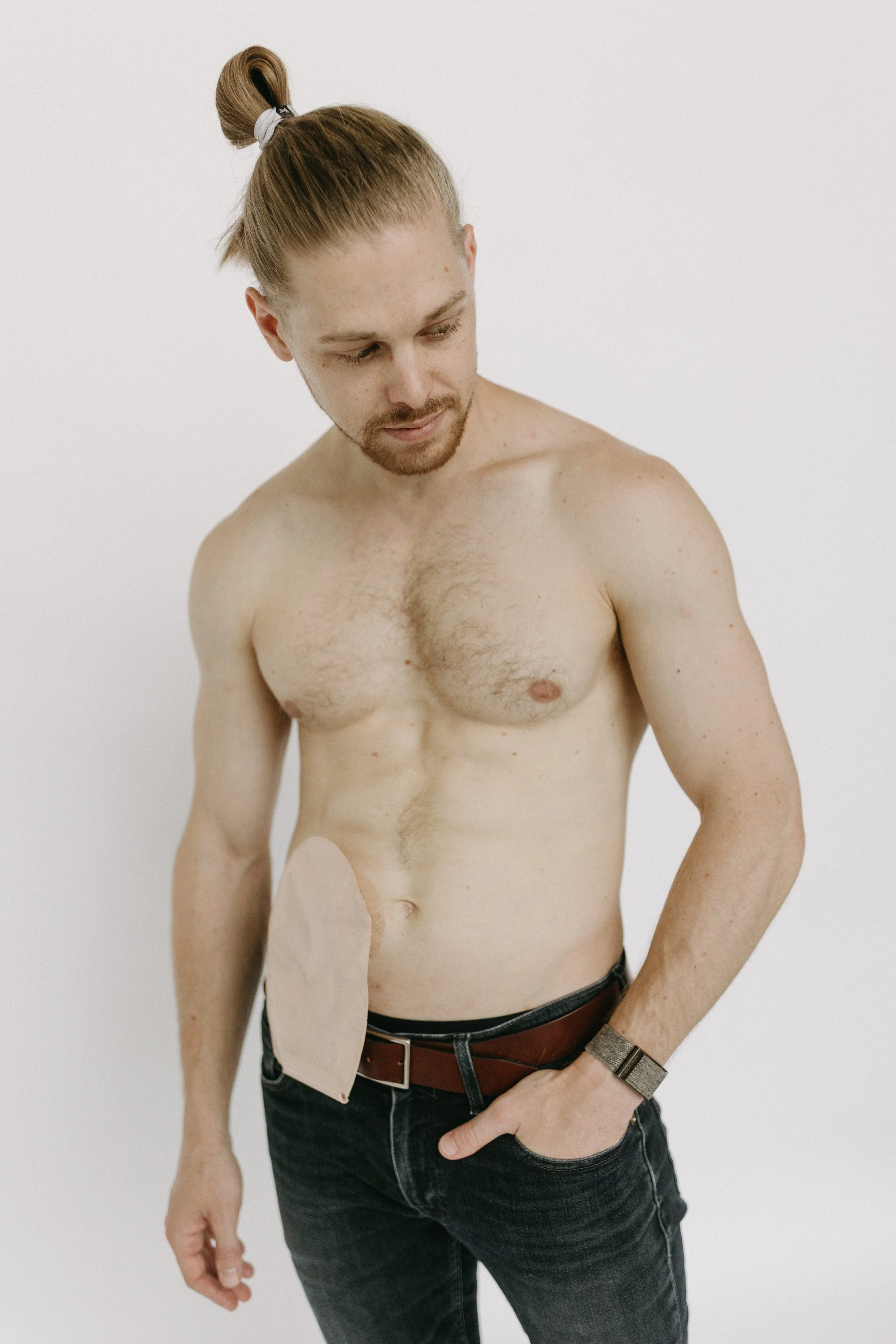 A shirtless man with long hair tied in a bun, looking down, with an ileostomy bag on his side and his hand in his pocket, standing against a plain white background.