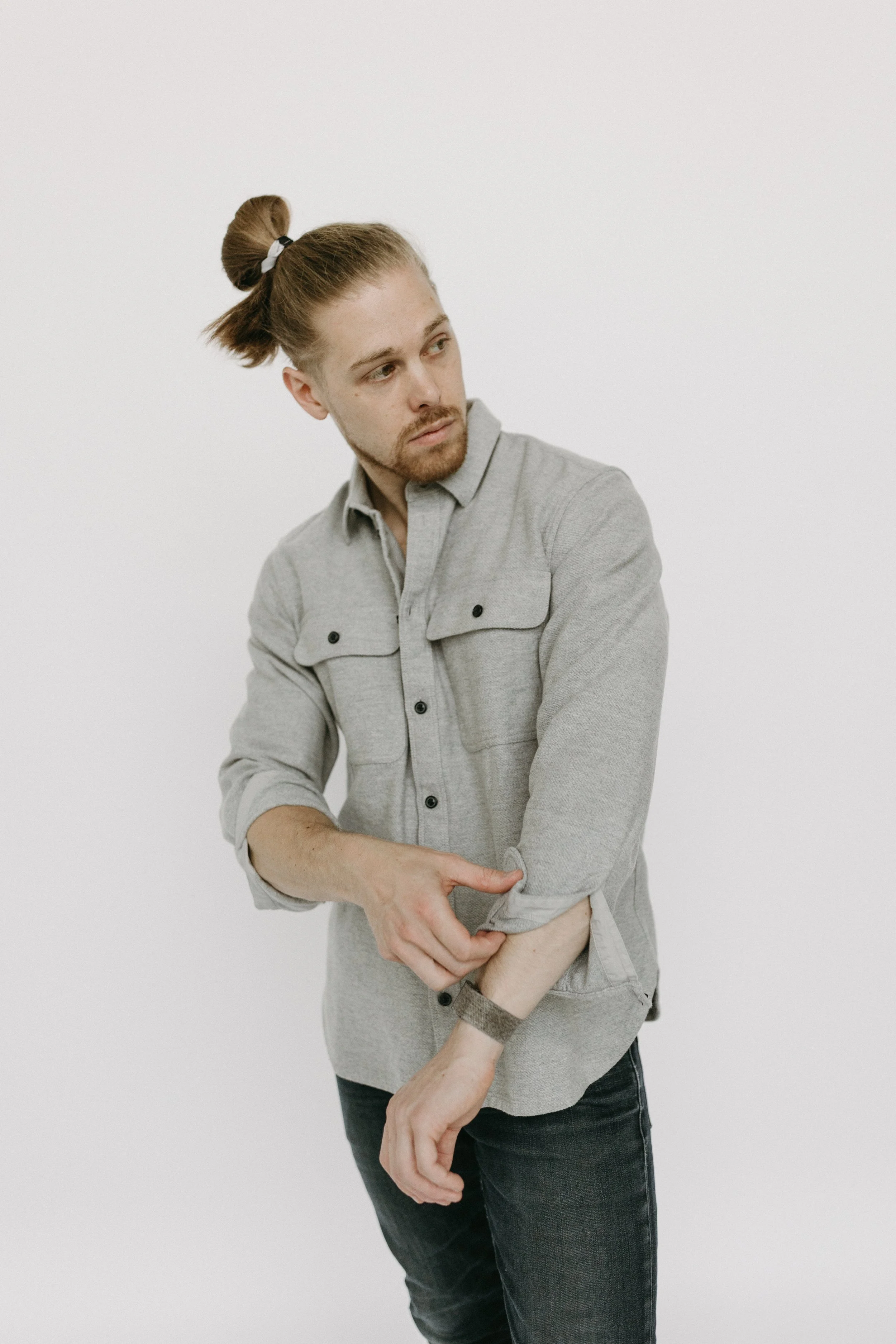 A man with a beard and tied-back hair is pulling up the sleeve of his gray shirt, wearing a black wristband, against a plain white background.