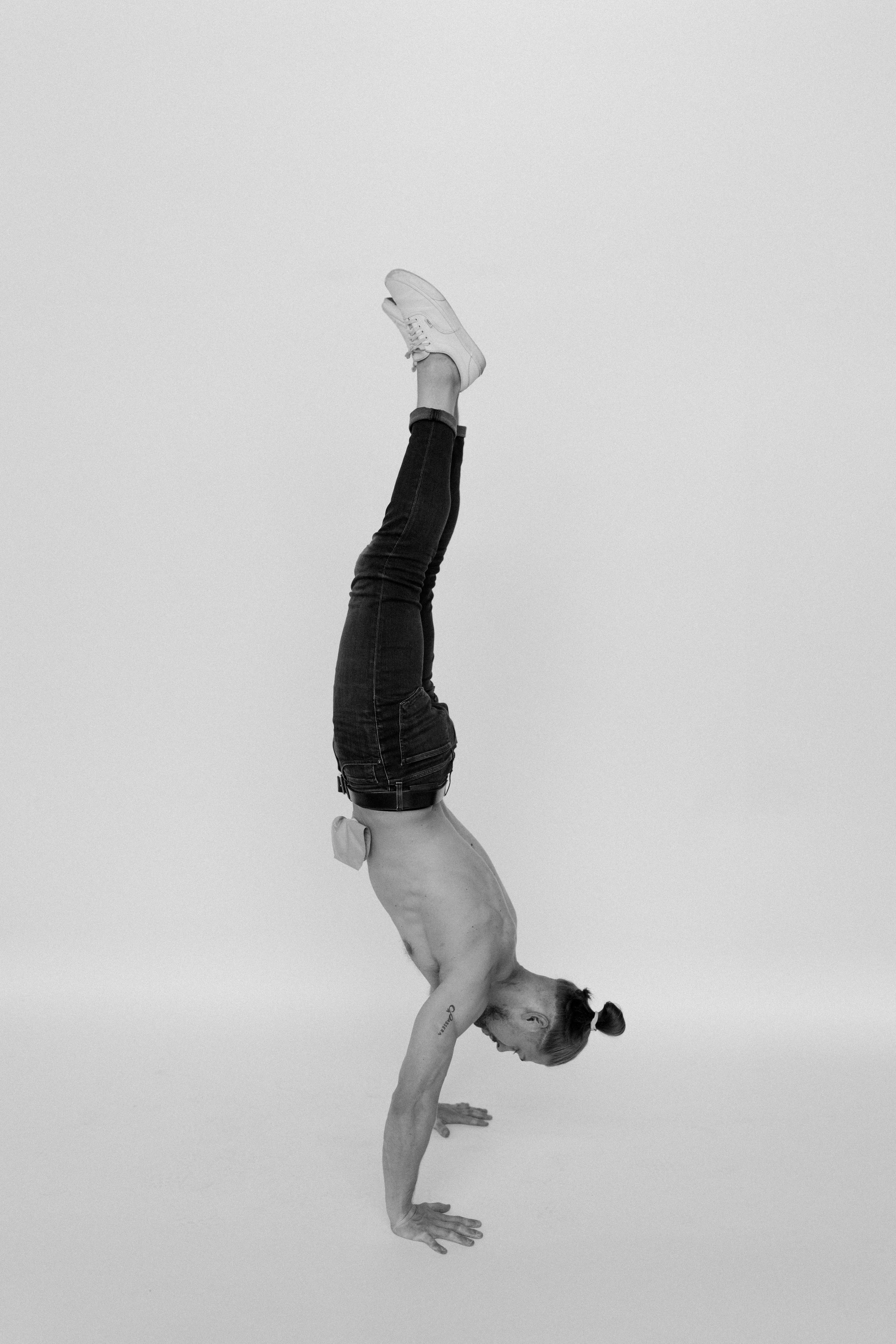A person performing a handstand with their legs extended upward, wearing jeans and sneakers, against a plain background.