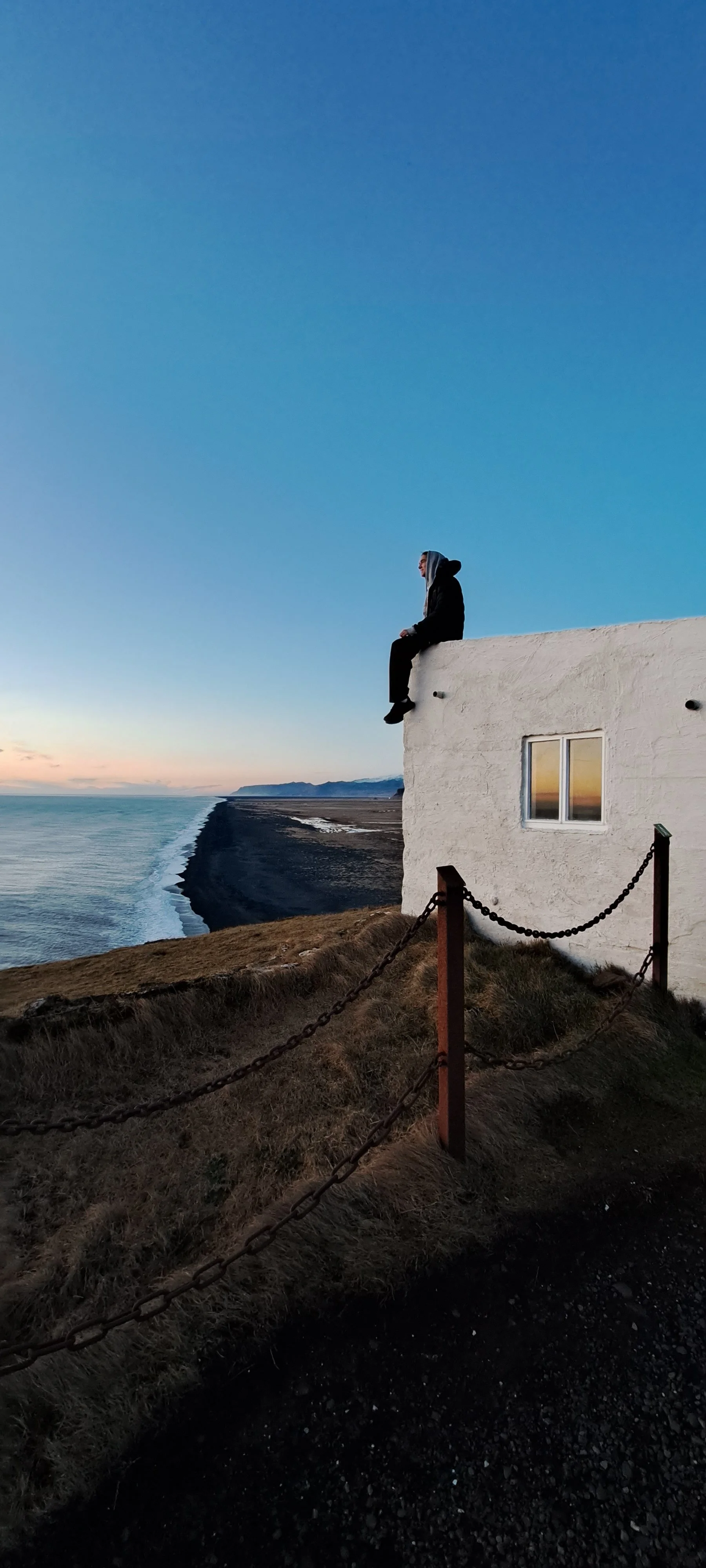 A person wearing a hoodie sitting on the edge of a white building overlooking the ocean at sunset.