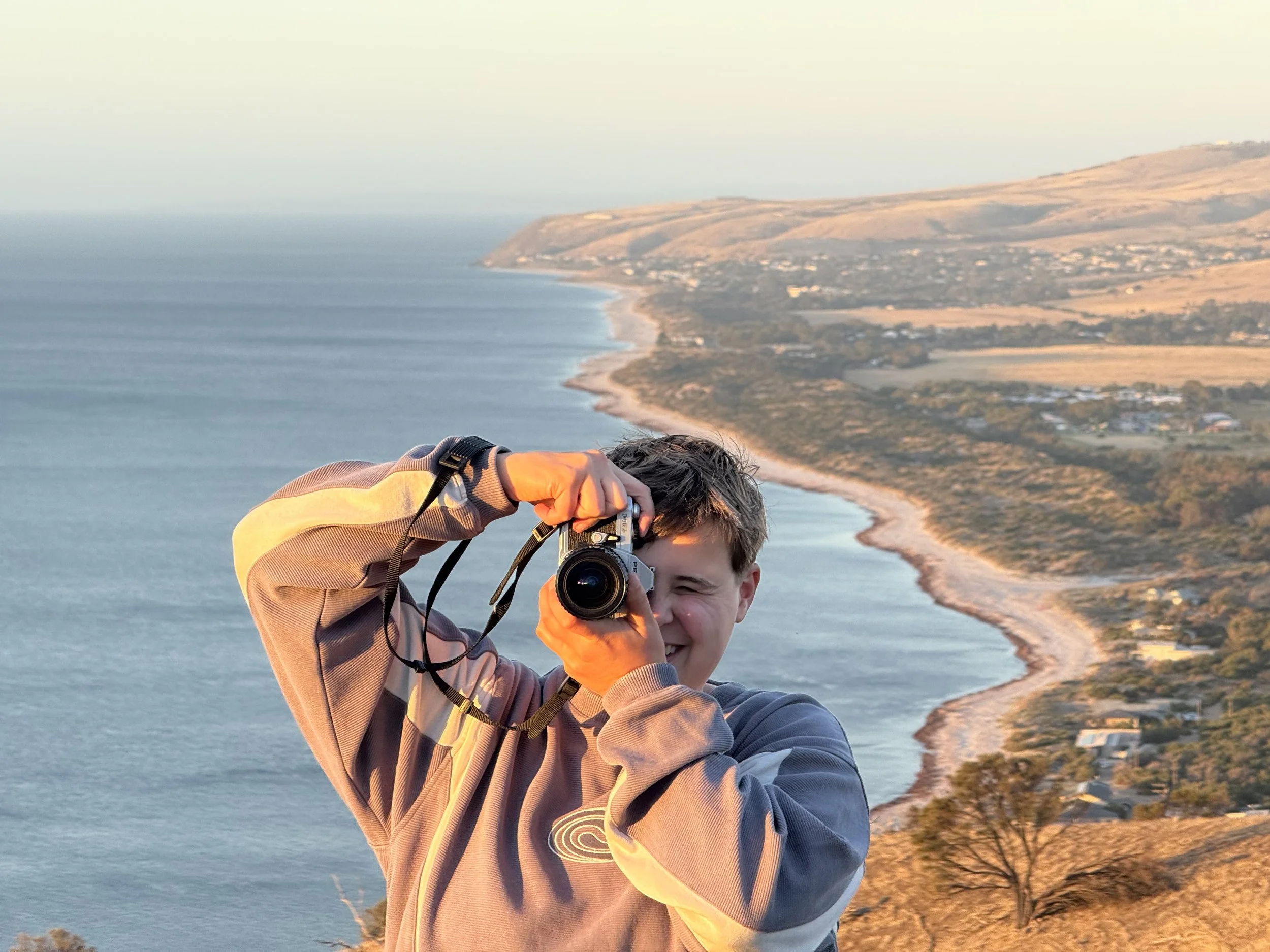 A young man taking a selfie with a camera on a scenic coastal cliff at sunset, overlooking the ocean and a distant shoreline with rolling hills and small buildings.