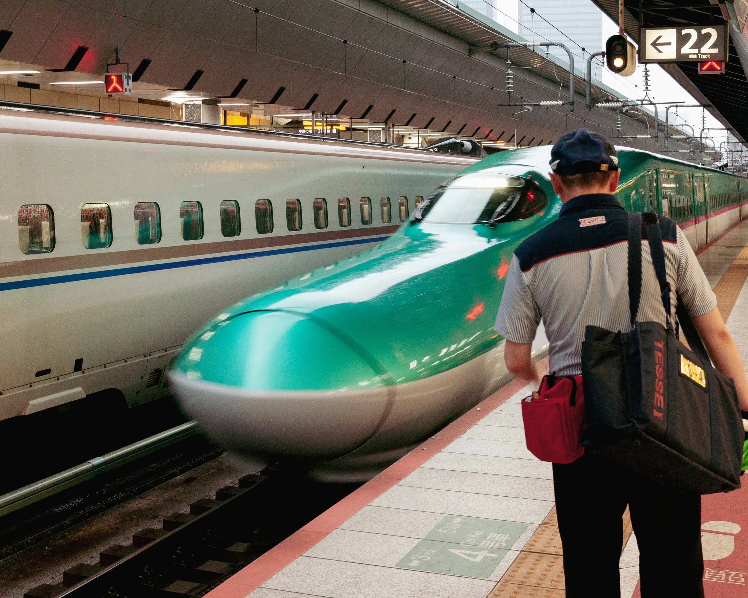 A man with a backpack is standing on a train platform, waiting to board a green high-speed train at a train station. The station has signs and a digital display overhead.
