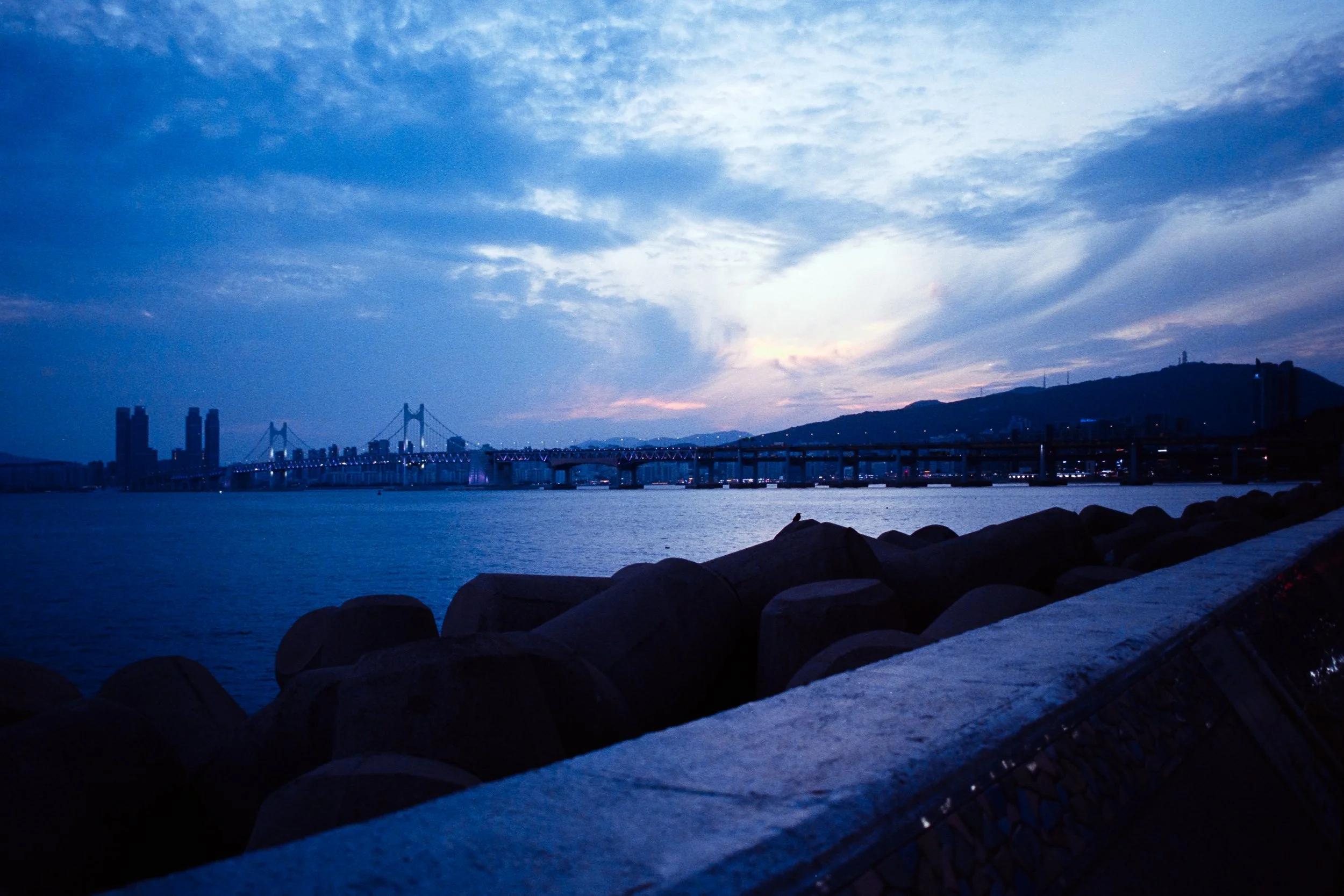 View of a bridge over water with city skyscrapers in the background and a mountaintop in the distance during twilight.