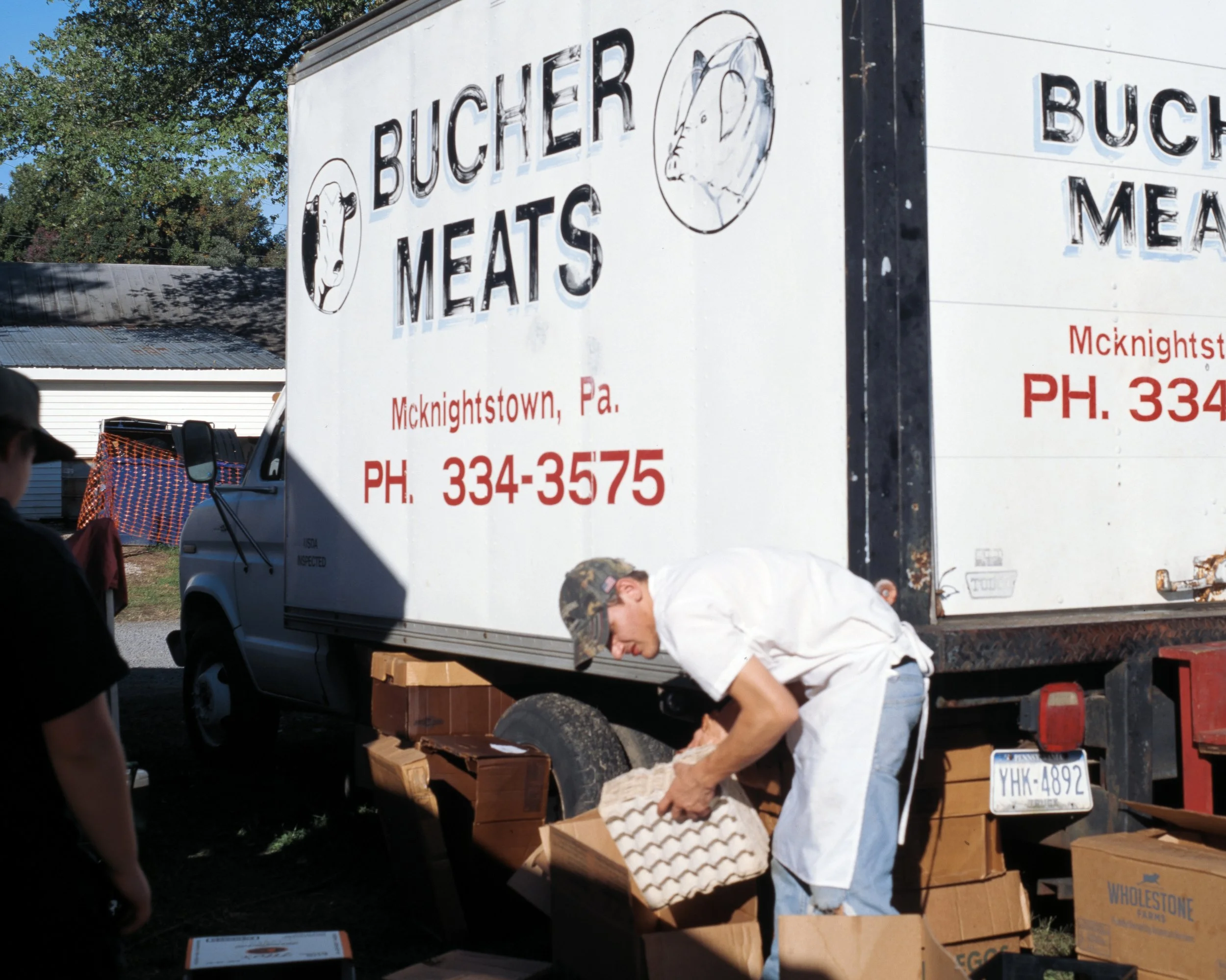 A man in a white shirt and camouflage cap is handling cardboard boxes near a white truck with 'Bucher Meats' and a logo of a pig's head on it, parked outdoors in daylight.