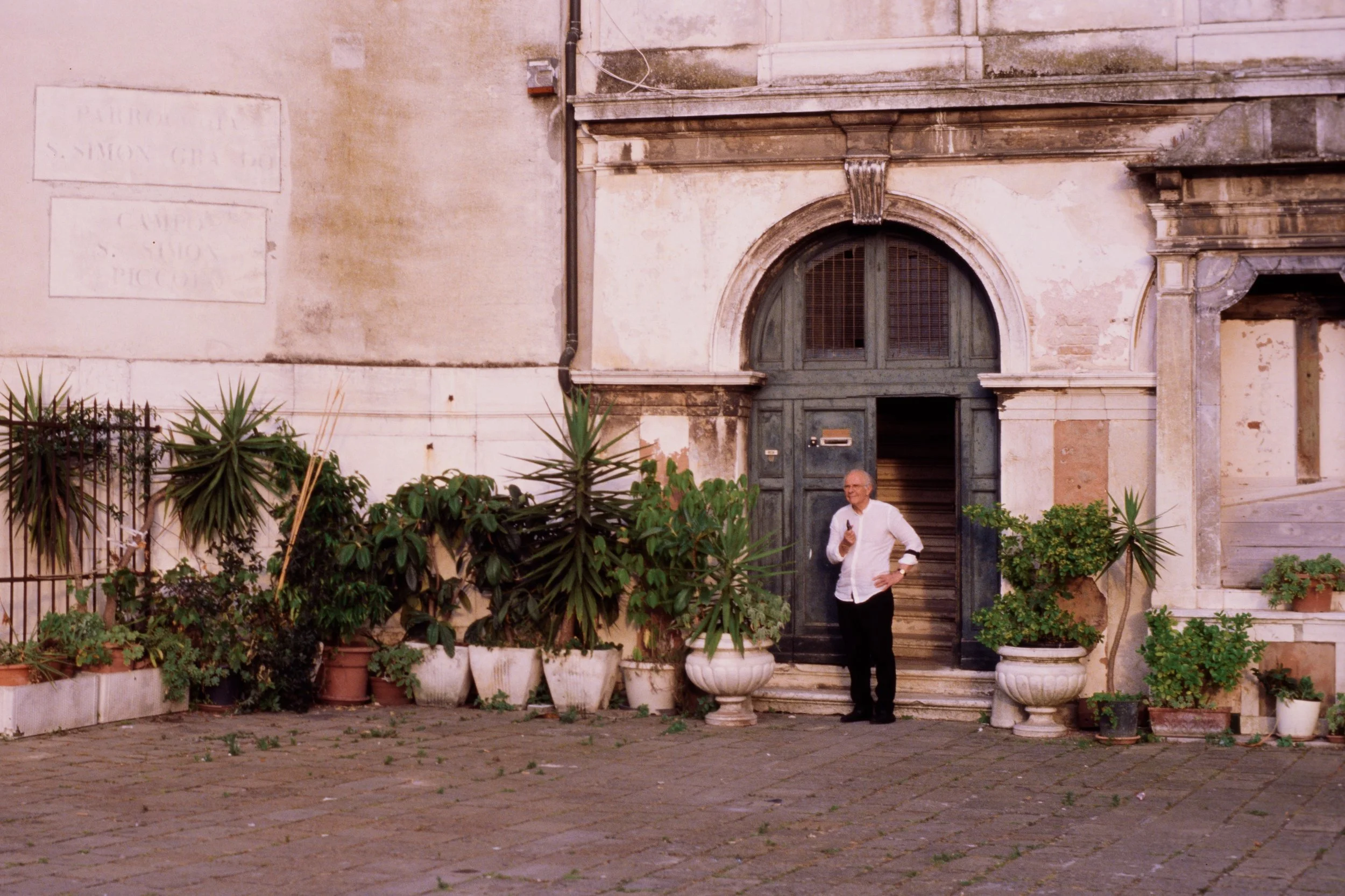An elderly man wearing a white shirt standing in front of a large weathered building with an arched green door and surrounded by potted plants.
