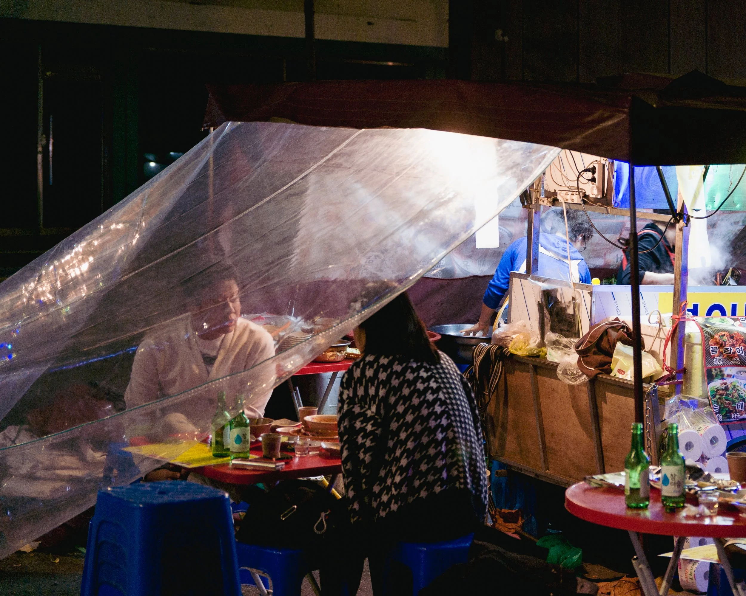Night scene of a street food stall with people preparing and eating, covered by a plastic sheet.