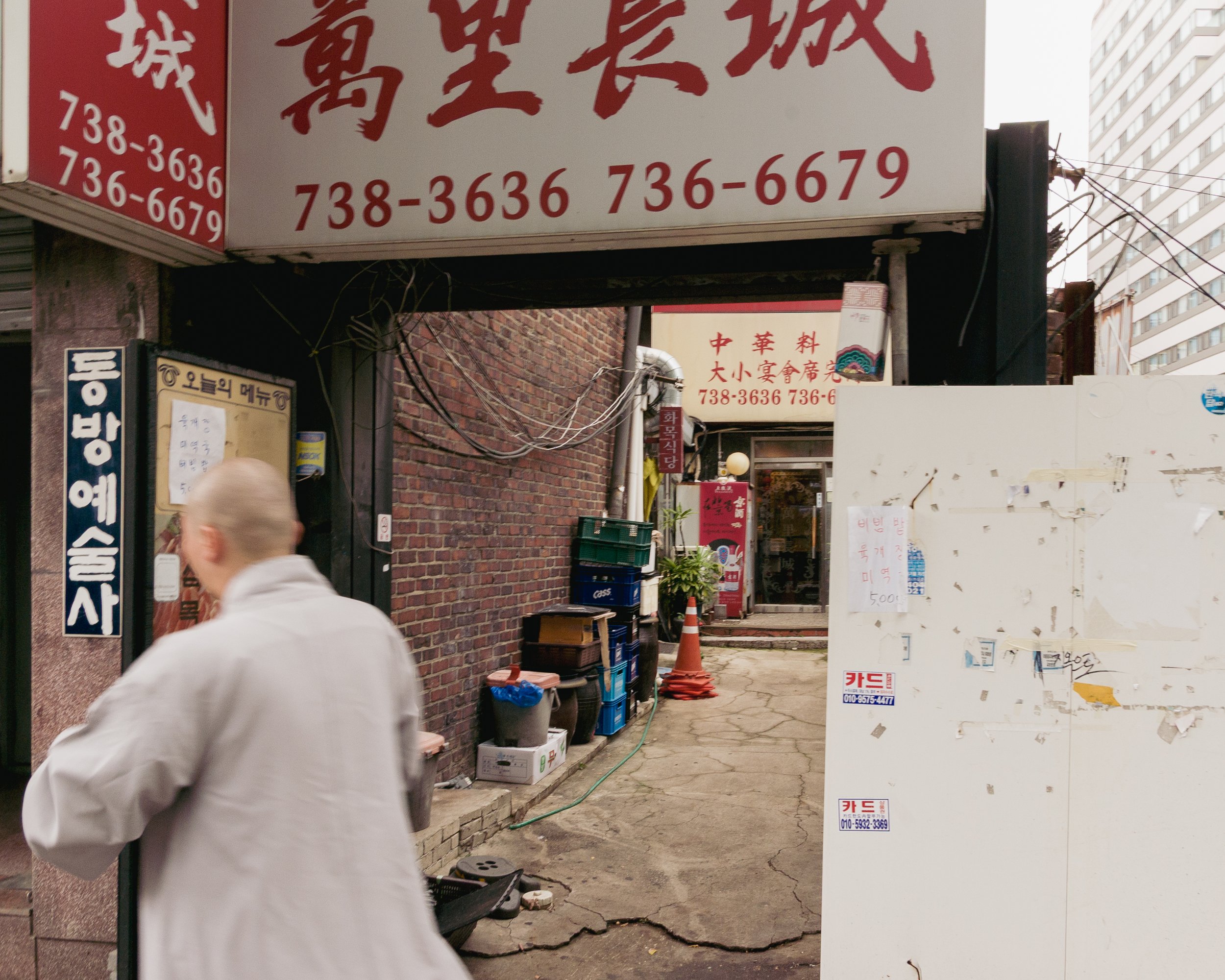 An alleyway in an urban setting with signs in Chinese and Korean, a person with a shaved head in a light-colored coat walking past, and various objects such as crates, potted plants, and traffic cones.