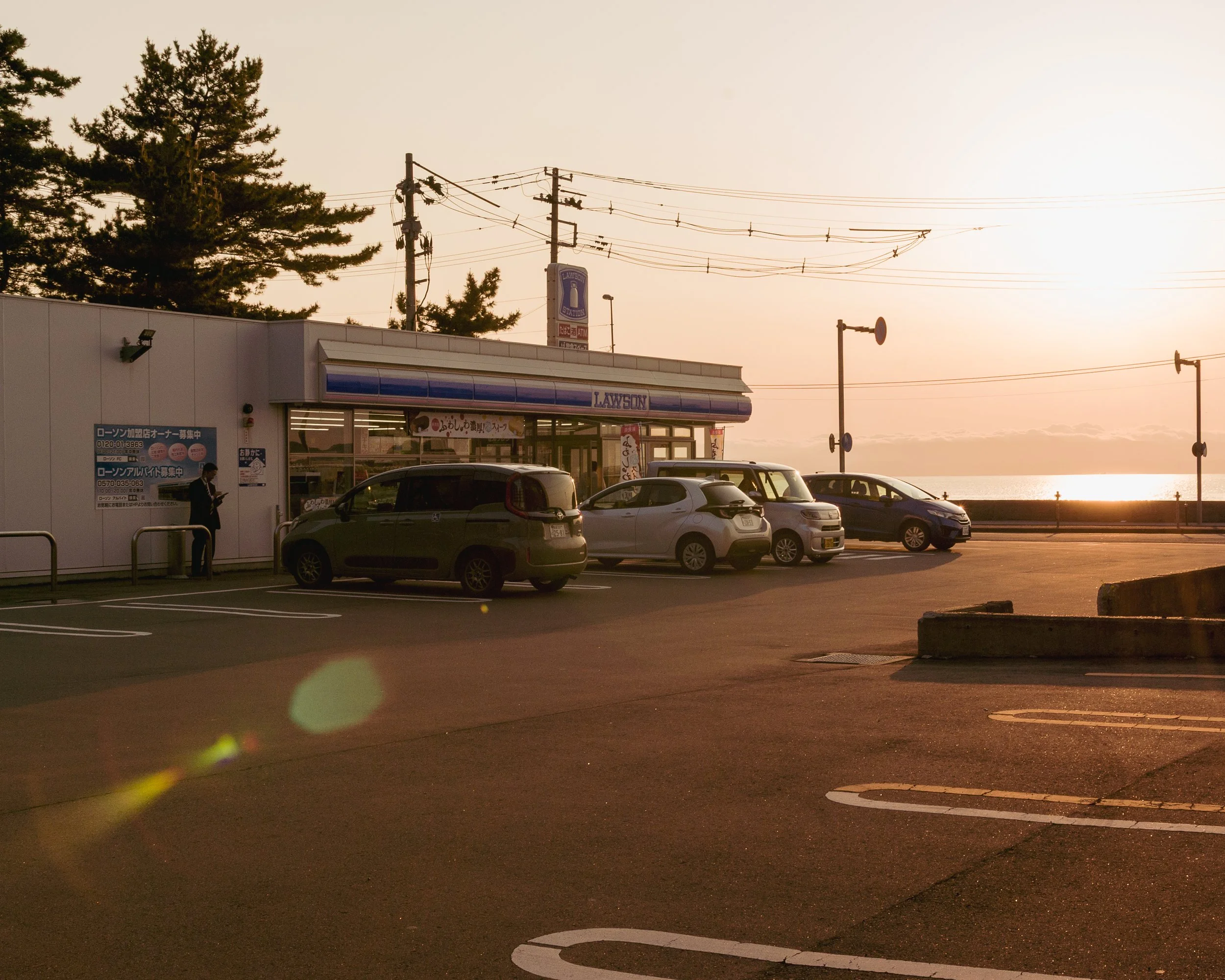 A parking lot with several parked cars in front of a Lawson convenience store at sunset, with trees and power lines in the background.
