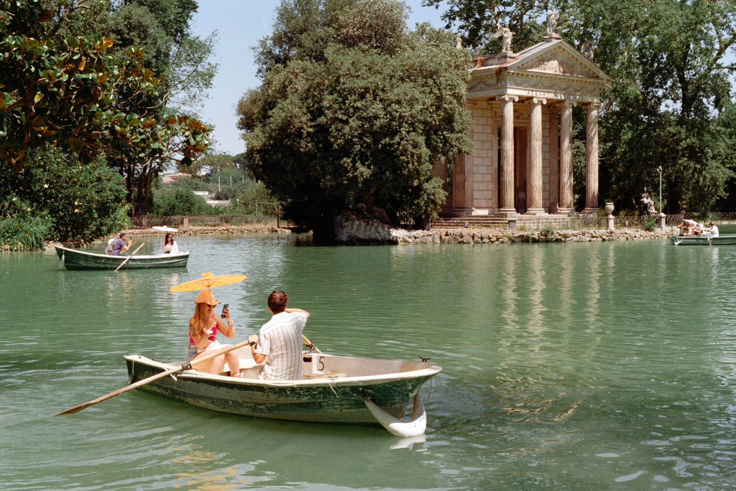 People boating on a lake with ancient Greek-style temple and greenery in the background.