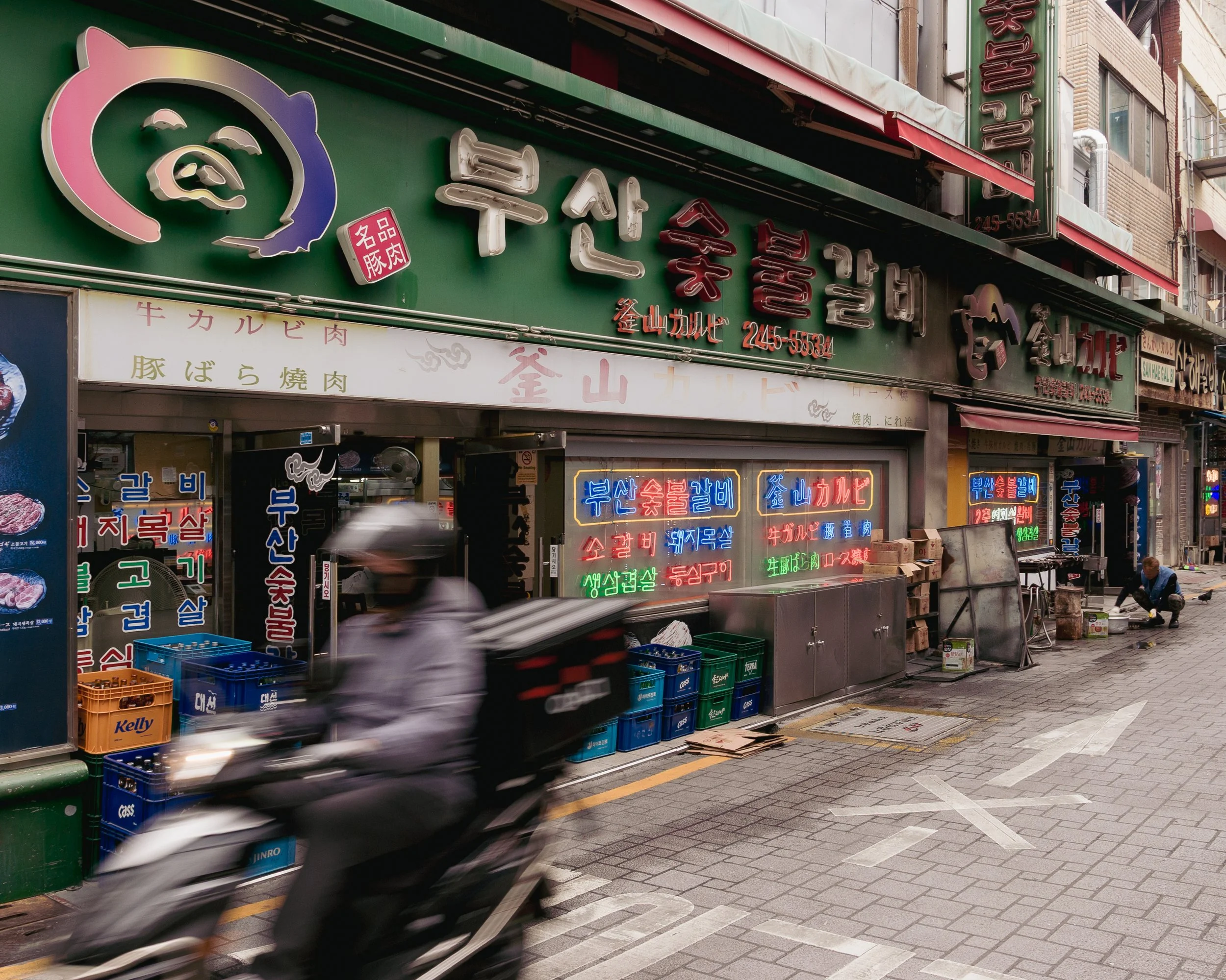 Street view of a Korean barbecue restaurant with neon signs in Korean, a motorcycle passing by, and a person crouching on the sidewalk.