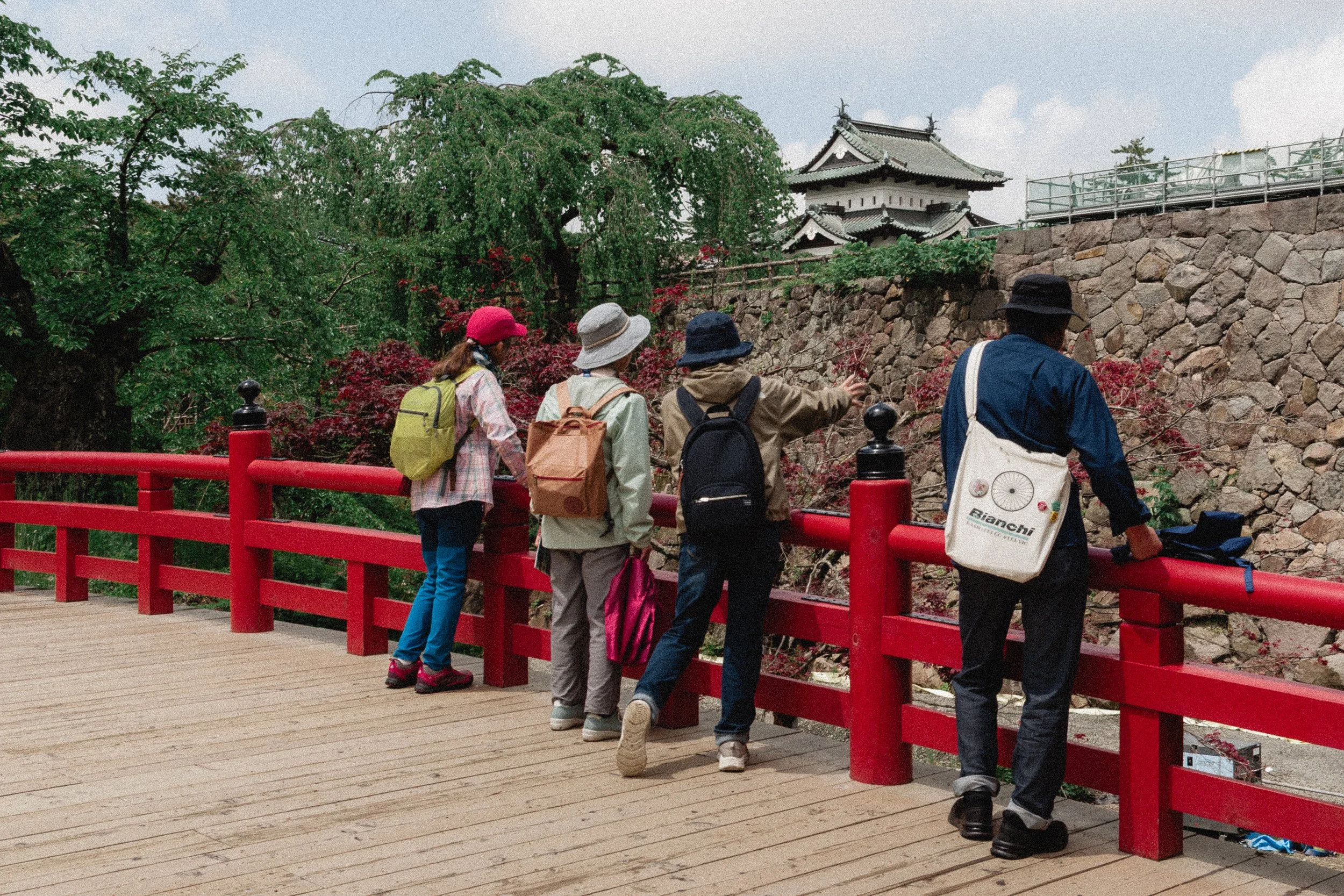 Group of people wearing backpacks and hats walking on a wooden bridge with a red railing, overlooking a stone wall and a traditional Japanese building, surrounded by green trees.