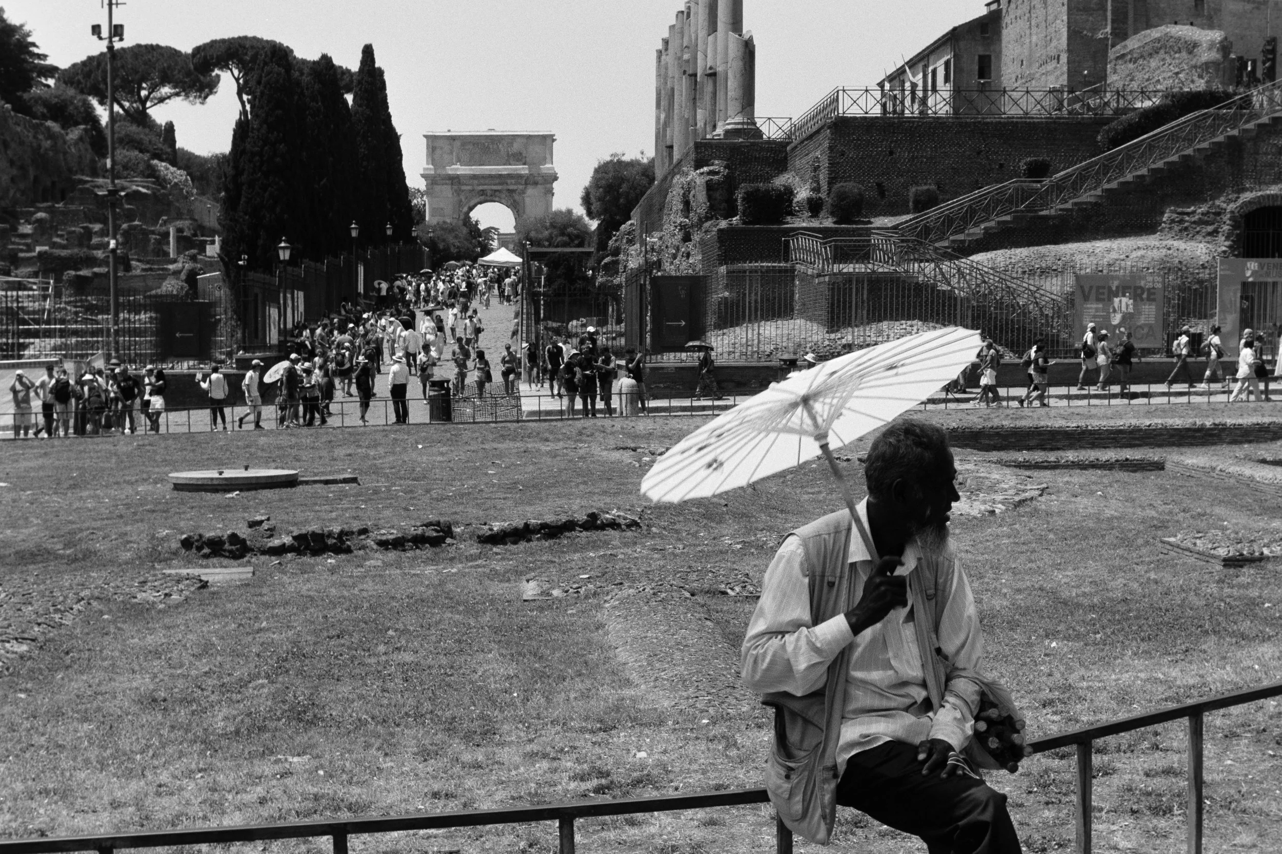 A black-and-white photo of a busy outdoor site with the Arc de Triomphe in the background. People walk along a pathway, some holding umbrellas. In the foreground, a man in a light-colored shirt and dark pants is sitting on a bench, holding an object 