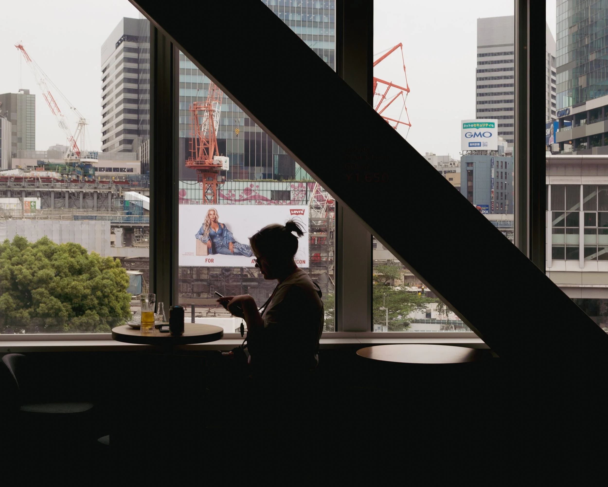 A woman sitting at a table with a drink, looking at her phone inside a modern building with large windows overlooking a cityscape with cranes and buildings.