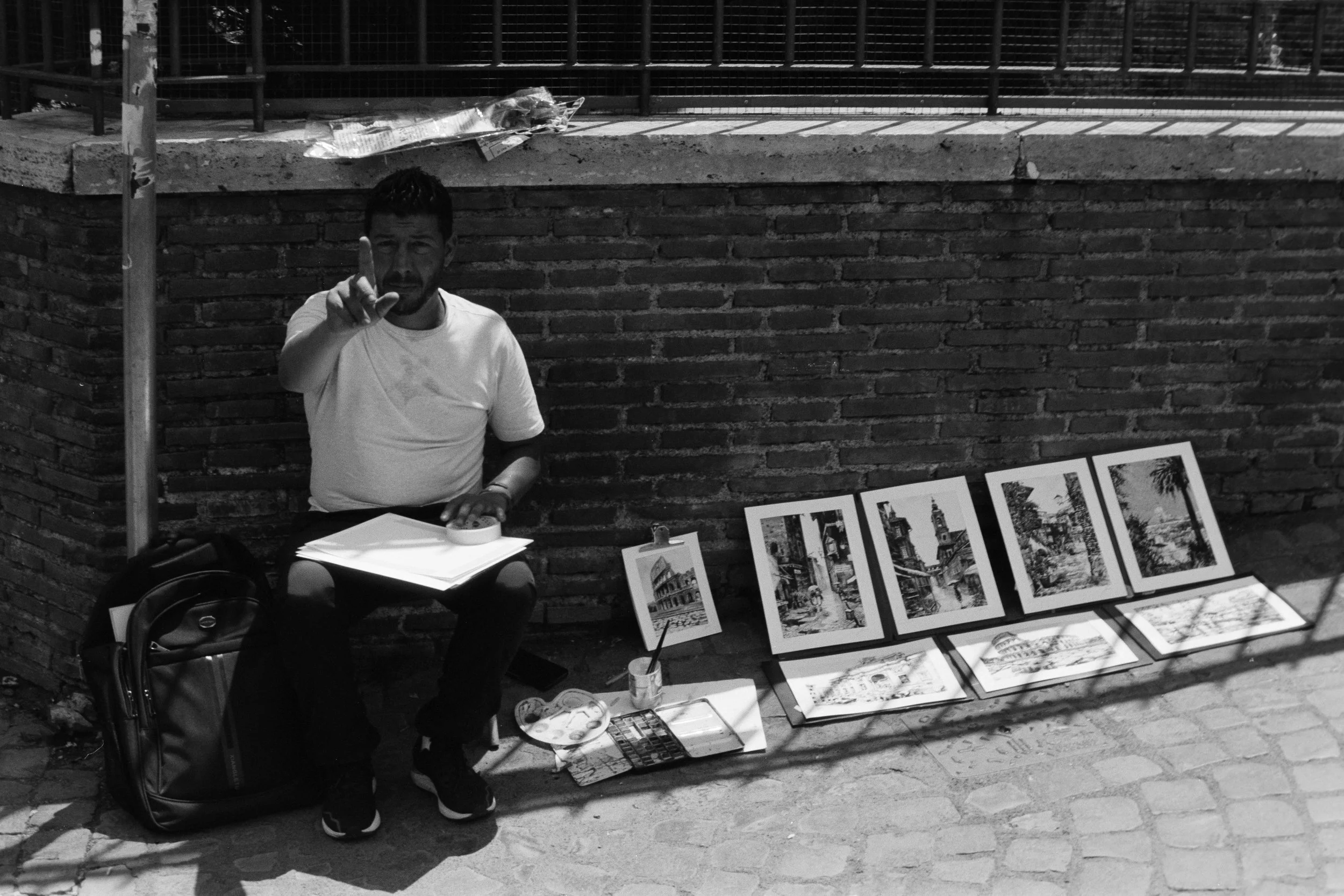 A street artist displaying prints of cityscape artwork at an outdoor market, sitting on a folding chair with a backpack and art supplies, with the art set arranged on the ground and leaning against a brick wall.