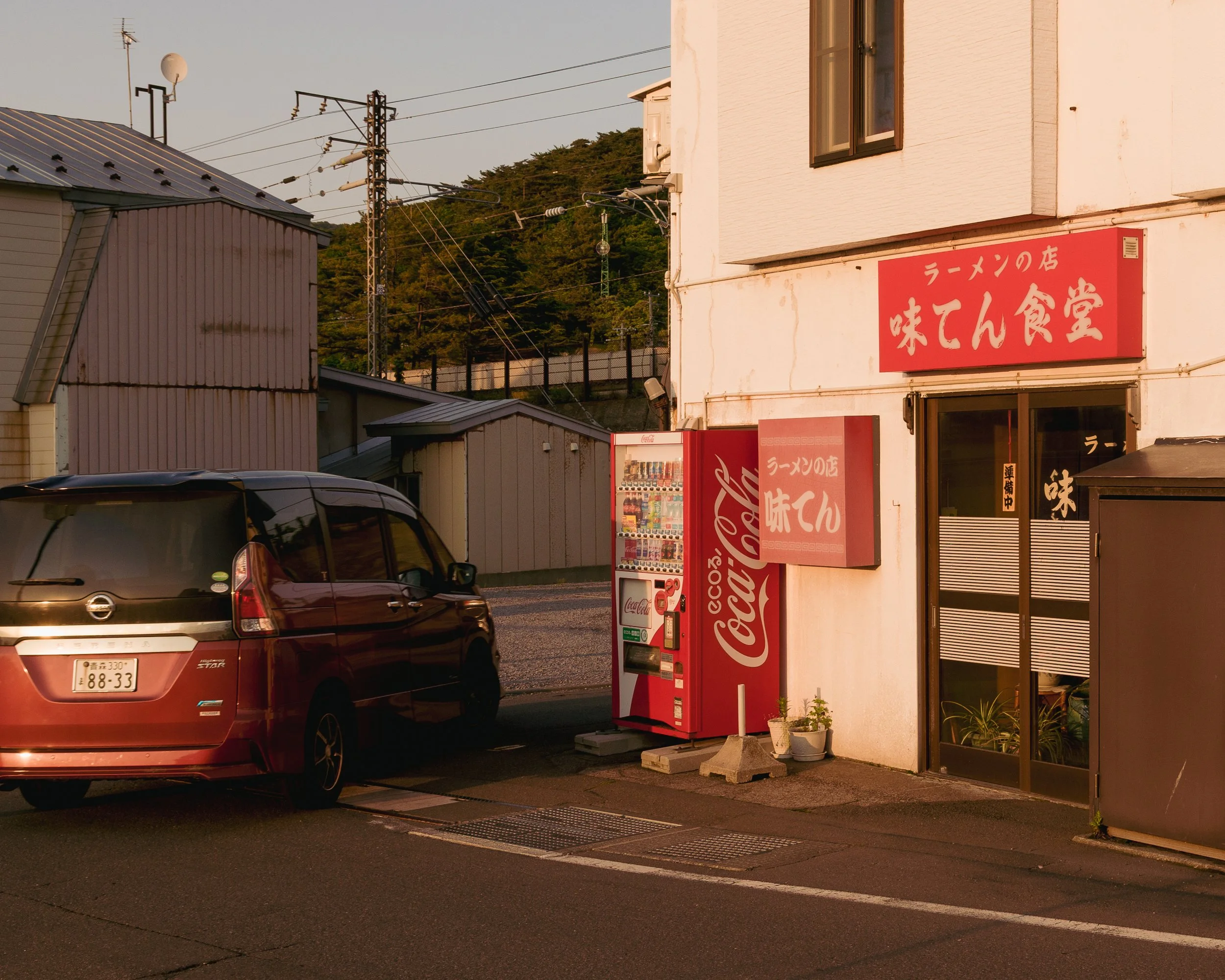 A red minivan parked in front of a small Japanese ramen restaurant during sunset, with a Coca-Cola vending machine next to the entrance and a red sign with white Japanese characters above the door.