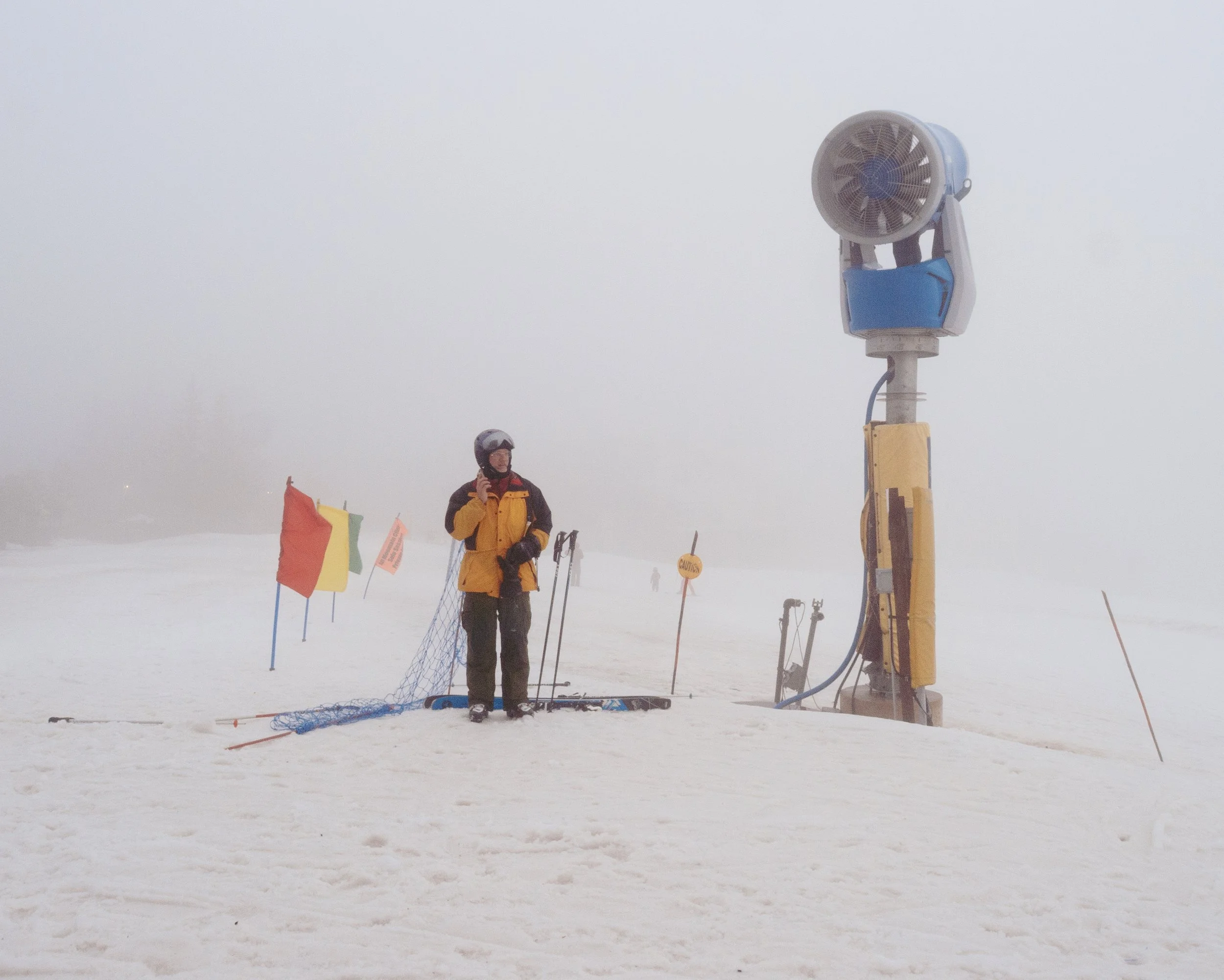 A person in winter gear standing on a snow-covered ski slope with a ski patrol station, ropes, and colorful flags in foggy conditions.