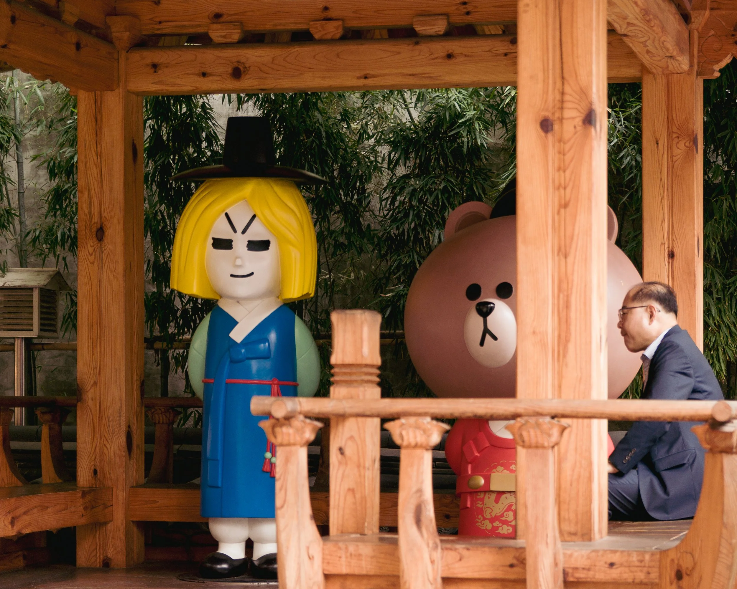 A man in a suit kneeling and talking to large cartoon character statues on a wooden structure. The characters include a woman in traditional Korean attire with a wide-brimmed hat and a bear in a red outfit with gold designs. The scene is set outdoors