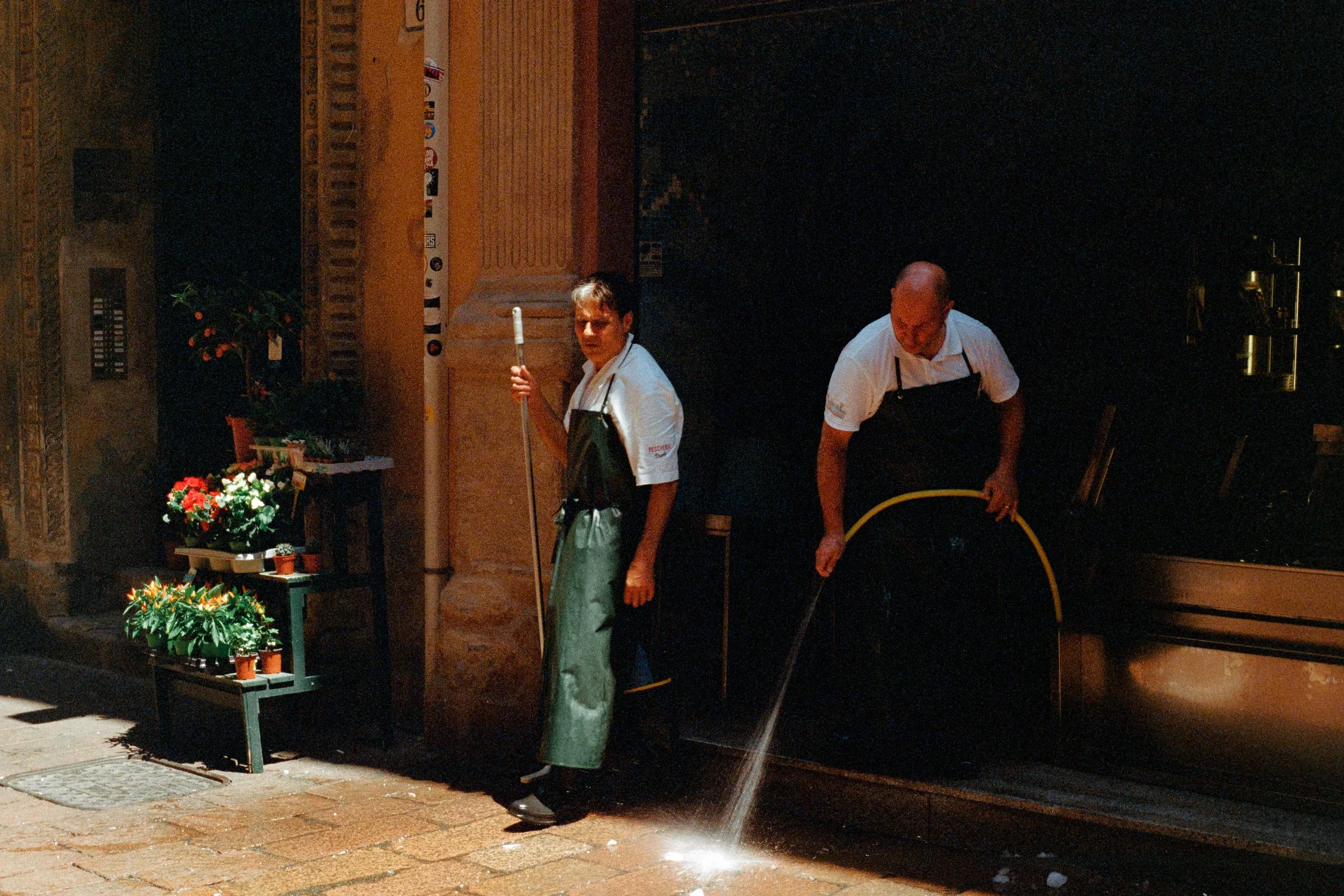 Two workers cleaning the street outside a flower shop at night. One is holding a mop, the other is handling a hose. Flower pots are visible on benches outside the shop.