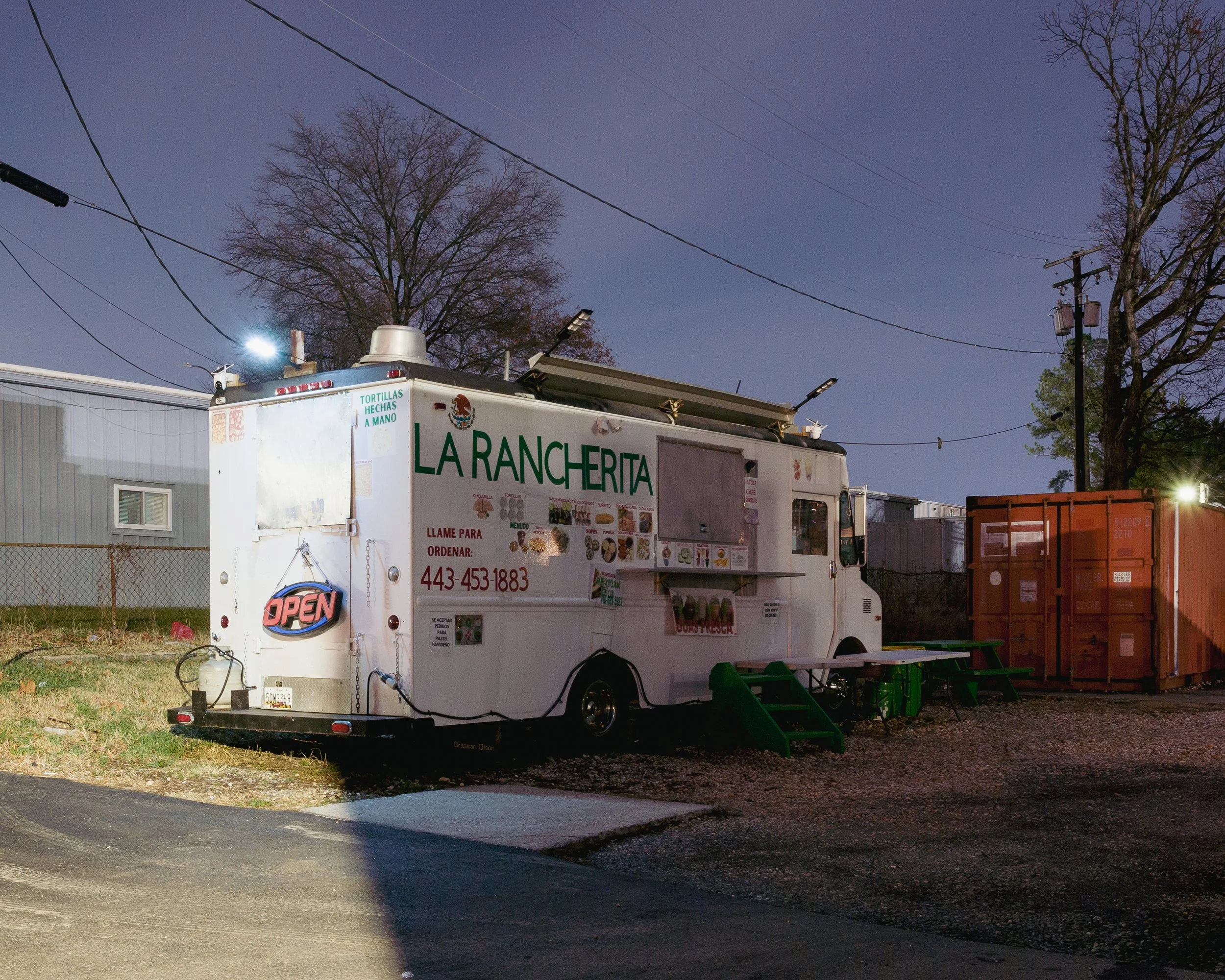 Food truck named 'La Rancherita' parked outdoors at dusk, serving Mexican food, with a small seating area and a bright 'Open' sign, surrounded by trees, power lines, and nearby containers.