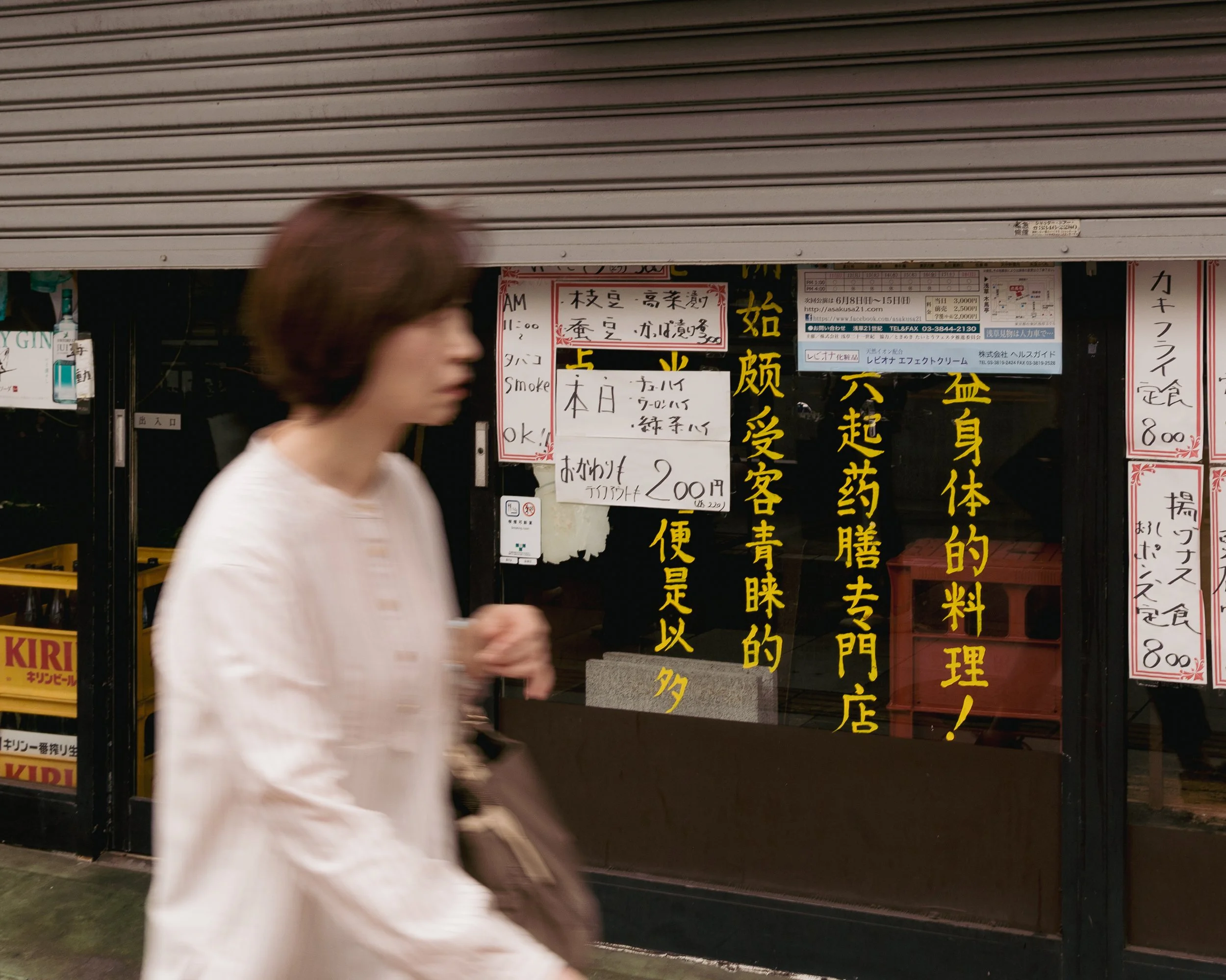 A woman with short brown hair wearing a white blouse walking past a restaurant window with Japanese signs and menus.