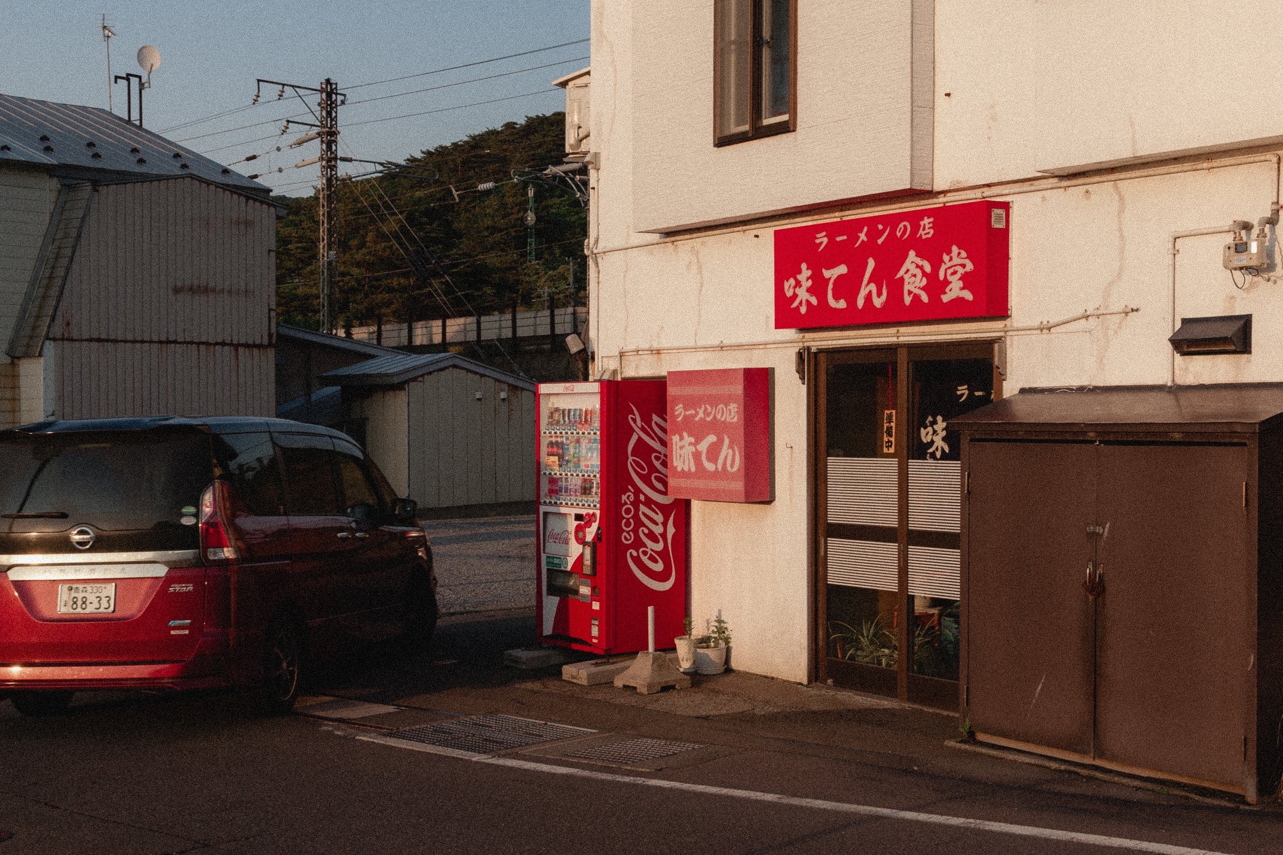 A red Coca-Cola vending machine outside a small eatery with a red sign in Japanese characters. The eatery is located next to a parking lot with a car. Power lines and a hill with trees are visible in the background.