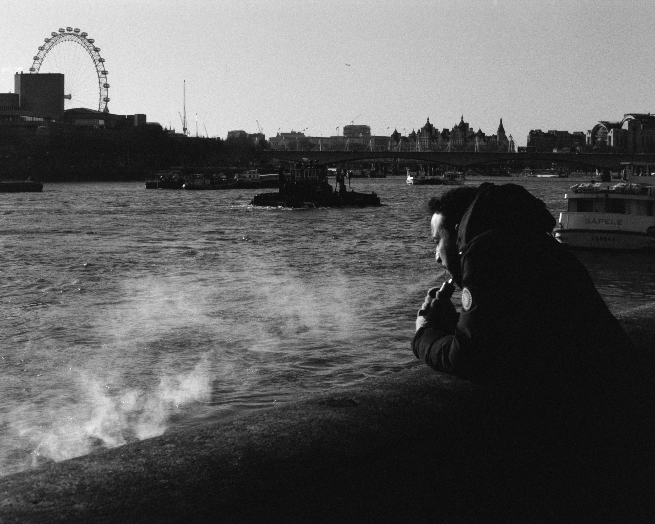 A man sitting by a river, smoking, with a city skyline and a Ferris wheel in the background.