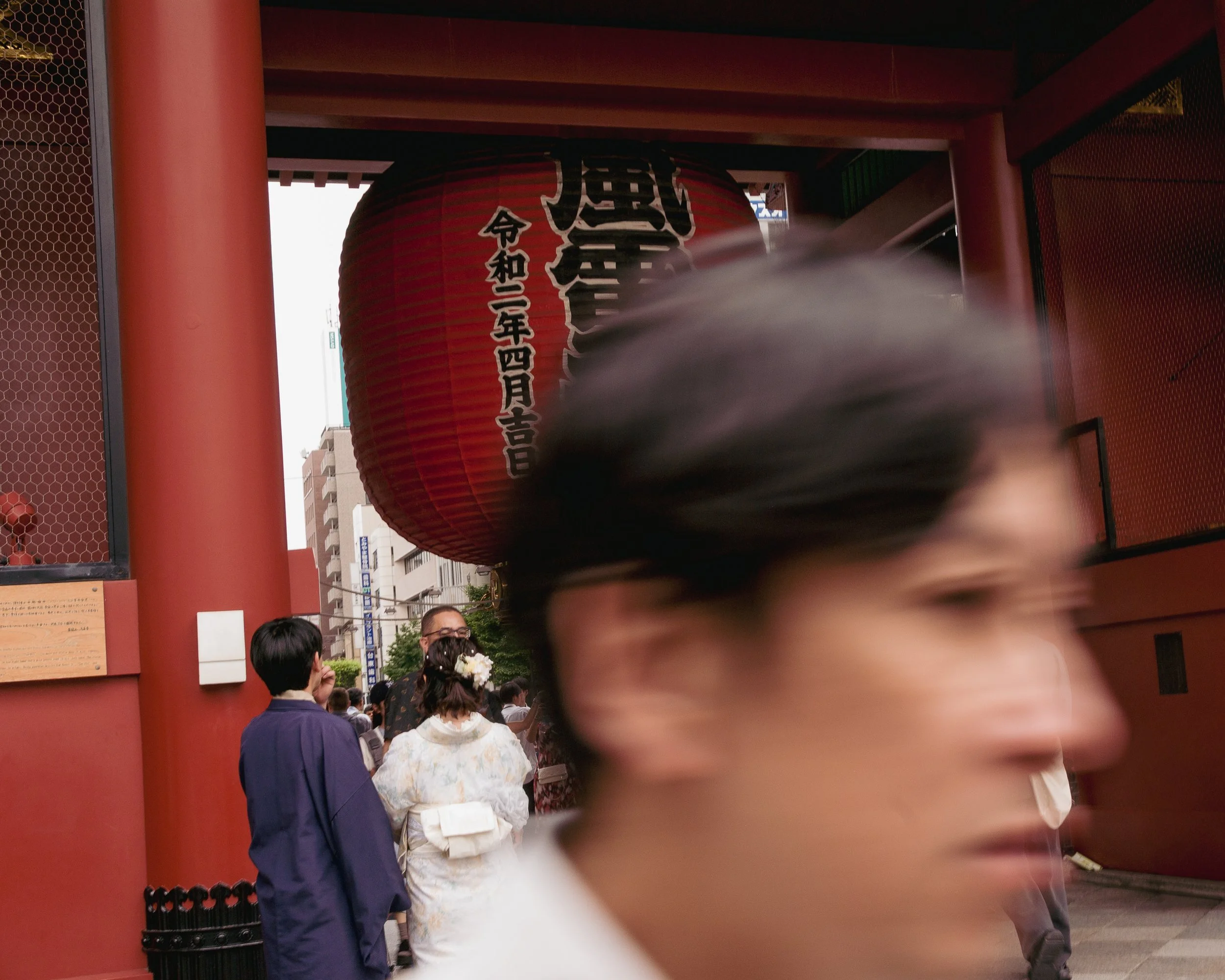 People gathered at the entrance of a Japanese shrine with a large red paper lantern hanging above, some dressed in traditional attire, with a blurred person in the foreground.