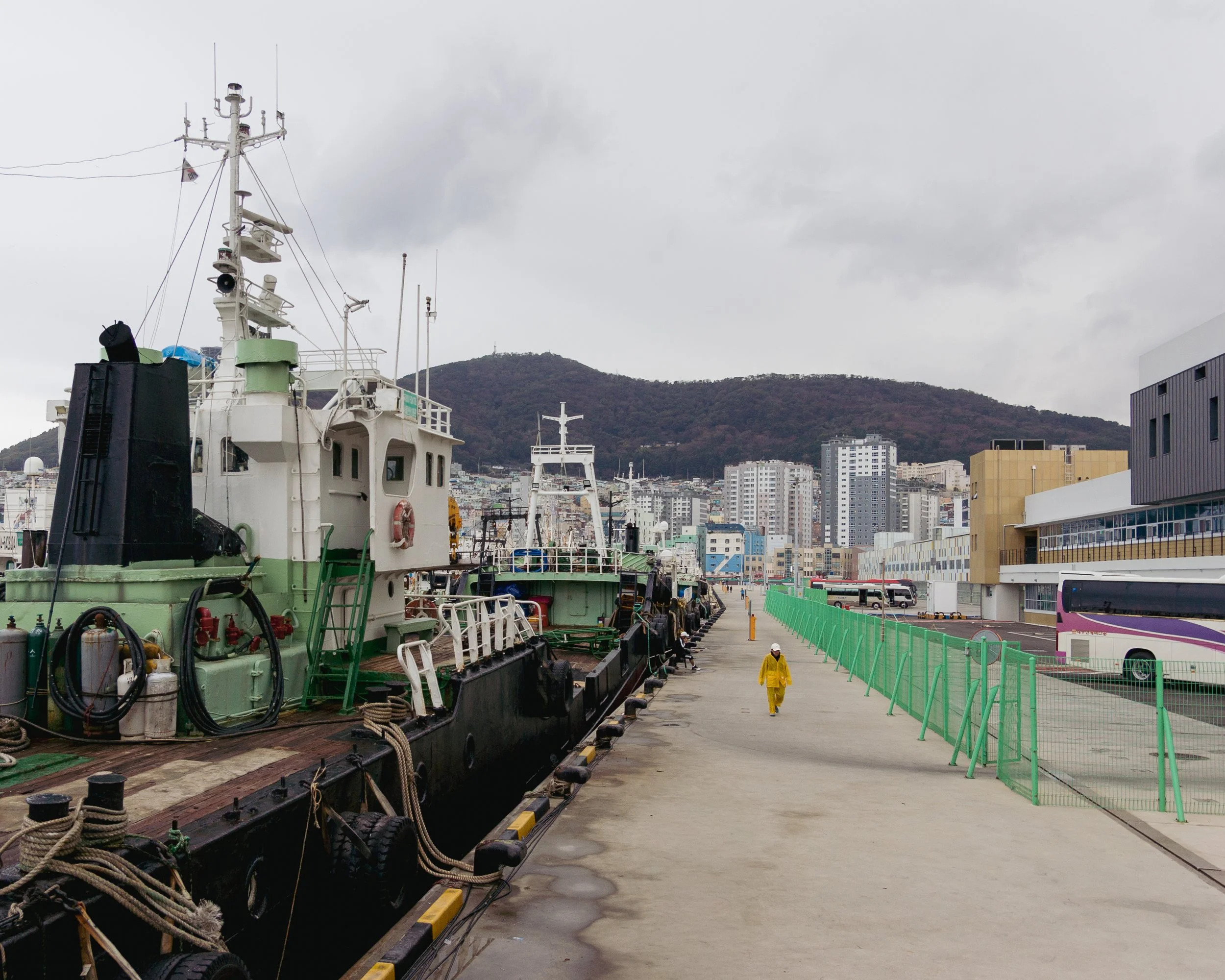 Dock area with ships docked on the left, city buildings in the background, and a person in a yellow raincoat walking on the sidewalk.