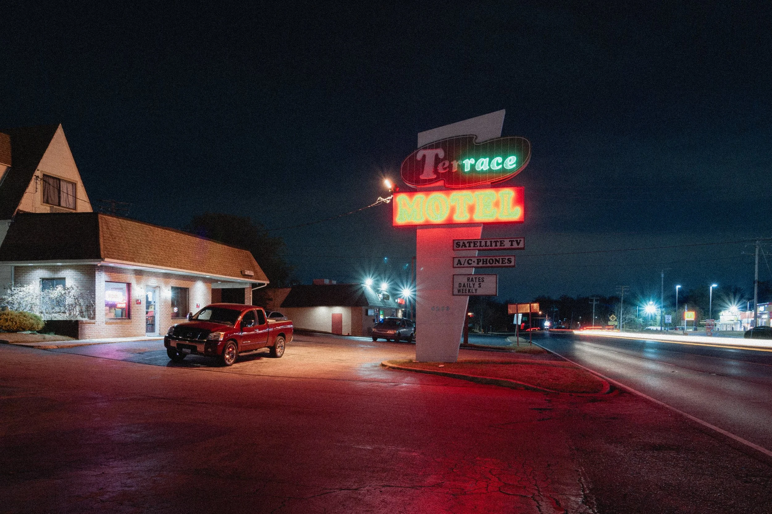 Nighttime scene of a Motel with neon signs reading 'Terrace' and 'Motel', along with smaller signs advertising satellite TV and air-conditioned rooms. The parking lot in front shows a red truck and other vehicles, with a building on the left and a da
