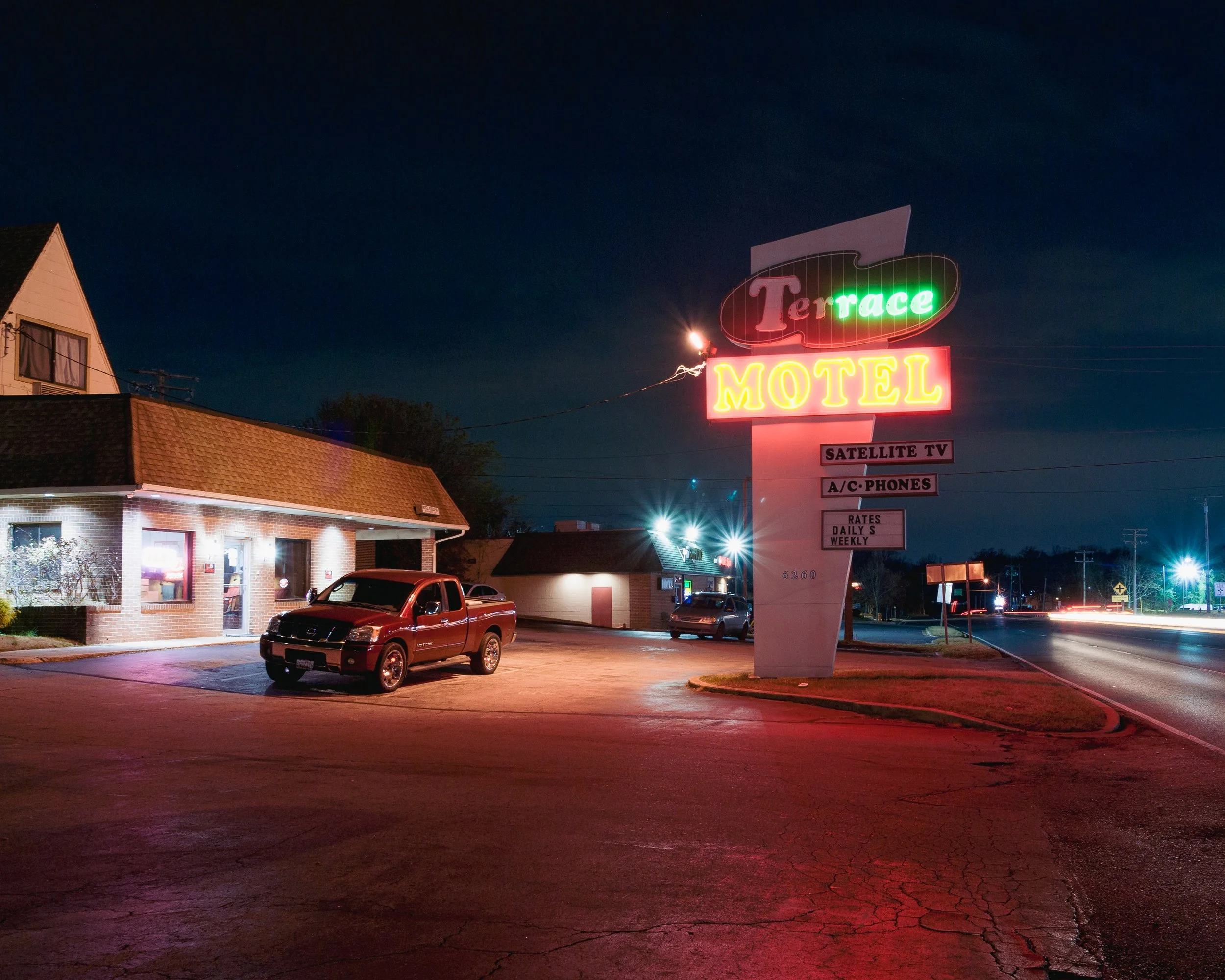 Nighttime scene of a small motel parking lot with a neon sign that reads 'Terrace Motel' and additional signs for satellite TV, A/C phones, and rates. There is a red pickup truck parked in front of a brick building with a lit window.