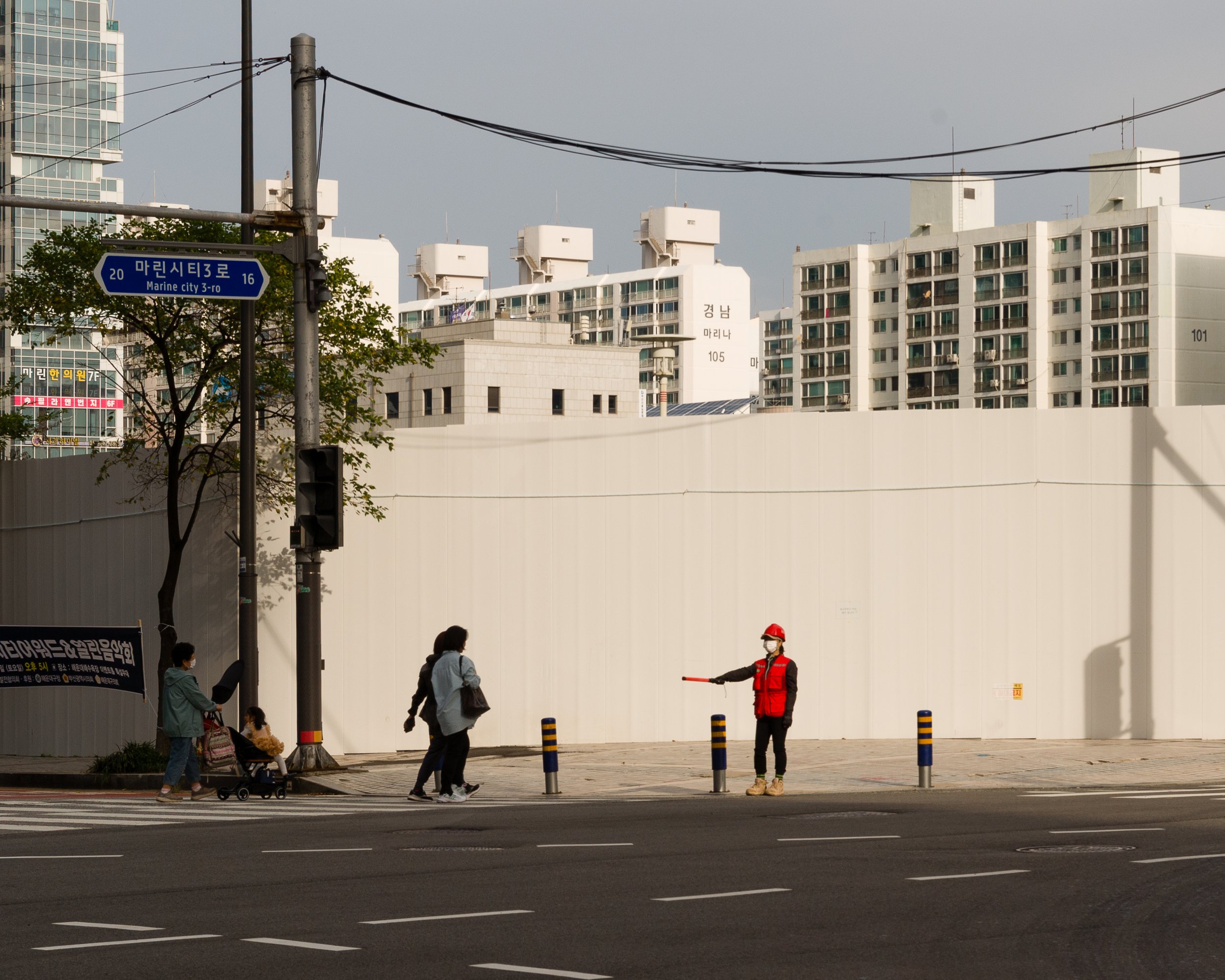 A person in a red vest and safety helmet directing pedestrian traffic at a crosswalk in an urban area with high-rise apartment buildings in the background.