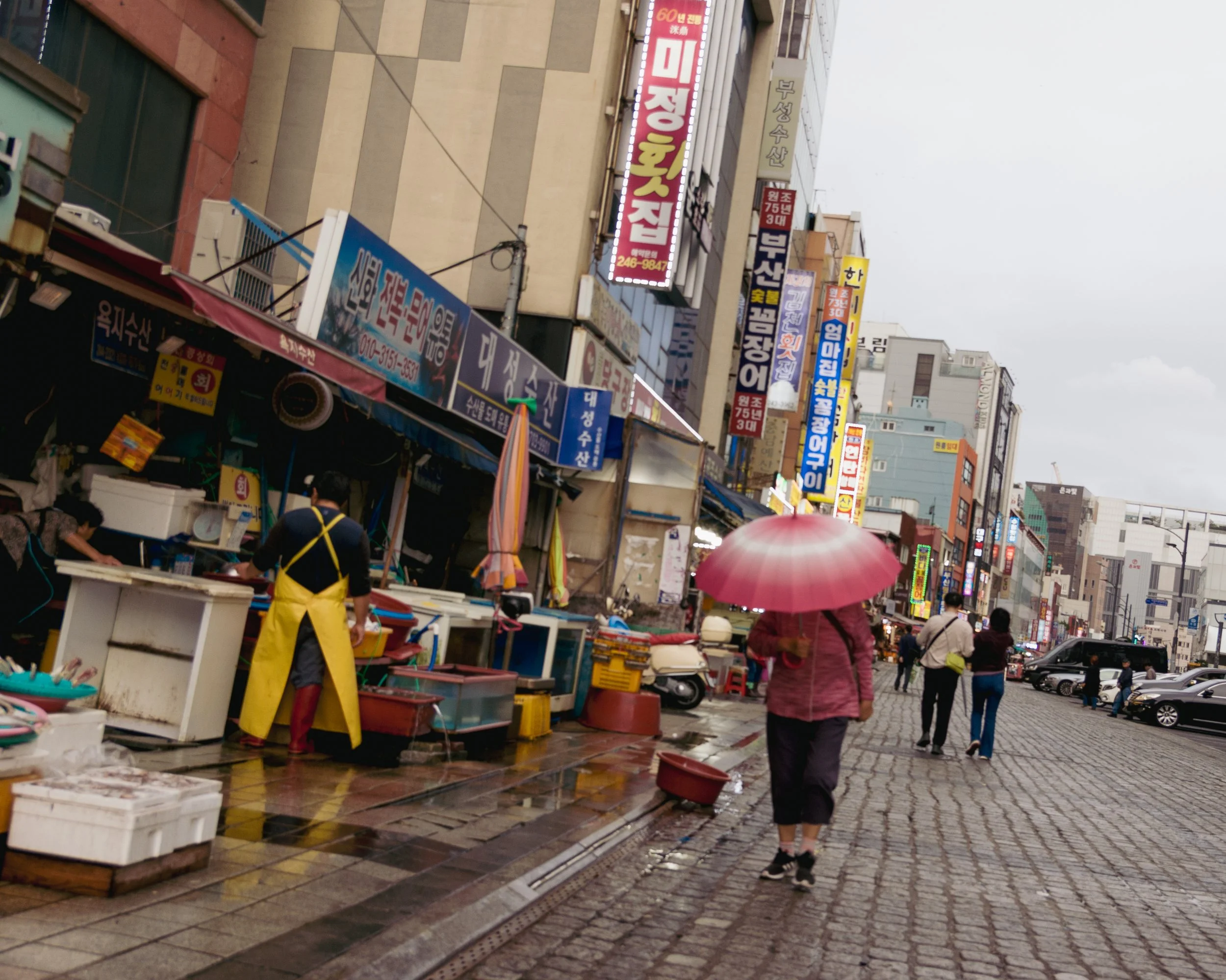 People walking on a city street on a rainy day, with signs in Korean on buildings and shops, one person holding a pink umbrella, and wet cobblestone pavement.