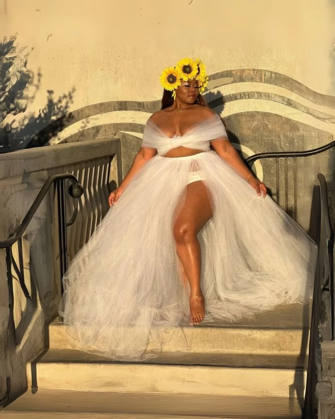 A woman wearing a sunflower crown and a sheer white off-shoulder dress with a high slit, standing on outdoor stairs against a textured wall with wave-like designs.