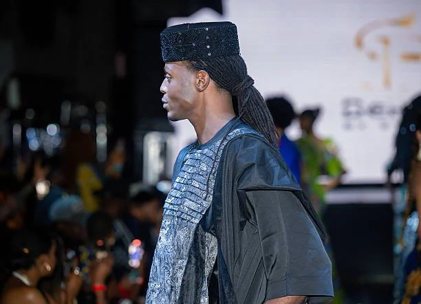 A man with long braided hair wearing a traditional black hat and an elaborate blue and black outfit, standing in a crowded indoor event with people and a screen in the background.