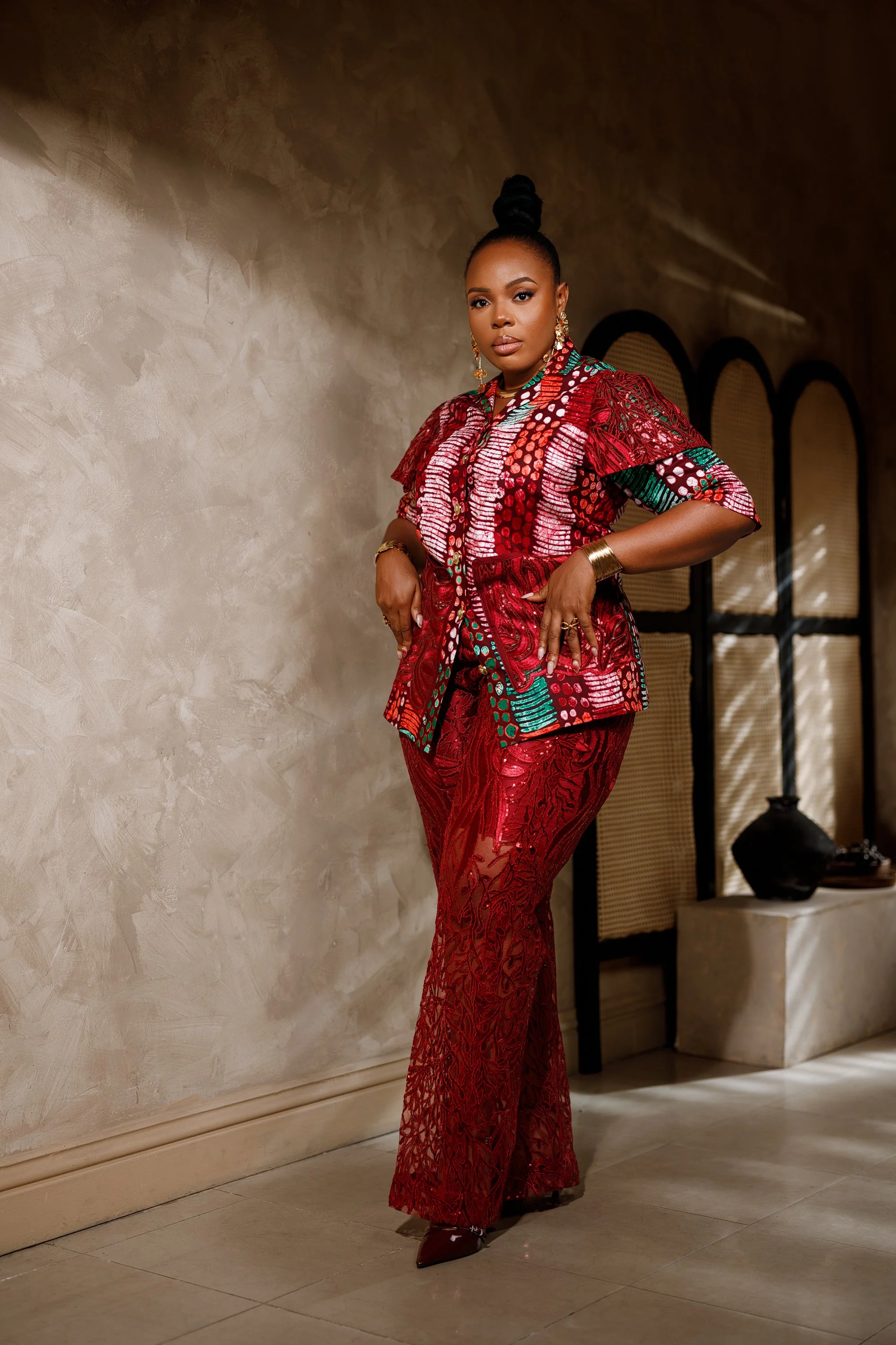 A woman dressed in a red, patterned, traditional African outfit with gold jewelry, standing indoors against a textured wall with arched windows in the background.