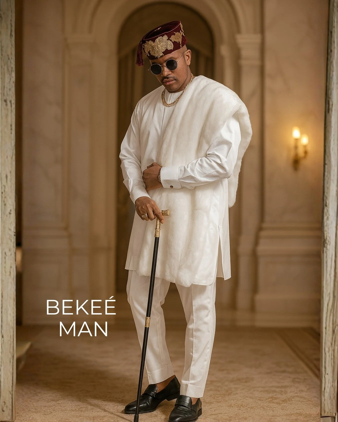 Man dressed in white traditional attire with a maroon and gold embroidered cap, sunglasses, gold jewelry, black loafers, holding a cane, standing in a luxurious interior with warm lighting.