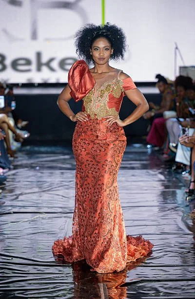 A woman walking down a runway in a red and gold lace evening gown with puffed sleeves and a train. She has curly black hair and hands on her hips.