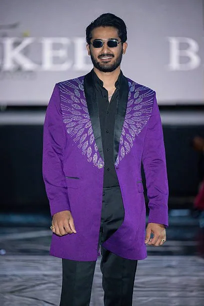A man walking on a runway, wearing sunglasses, a purple blazer with a feather pattern on the shoulders, a black shirt, and black pants.