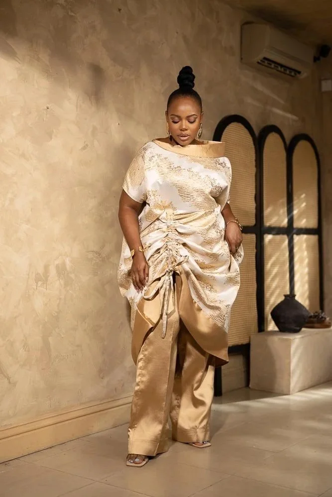 A woman dressed in a matching gold and beige shiny outfit with loose pants and a ruched top, standing indoors against a textured beige wall, with her hair styled in a high bun and wearing large hoop earrings.