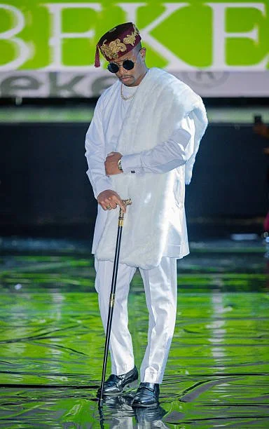 A man dressed in all white traditional attire with a colorful headwrap, sunglasses, and jewelry, standing on a shiny green stage and holding a cane.