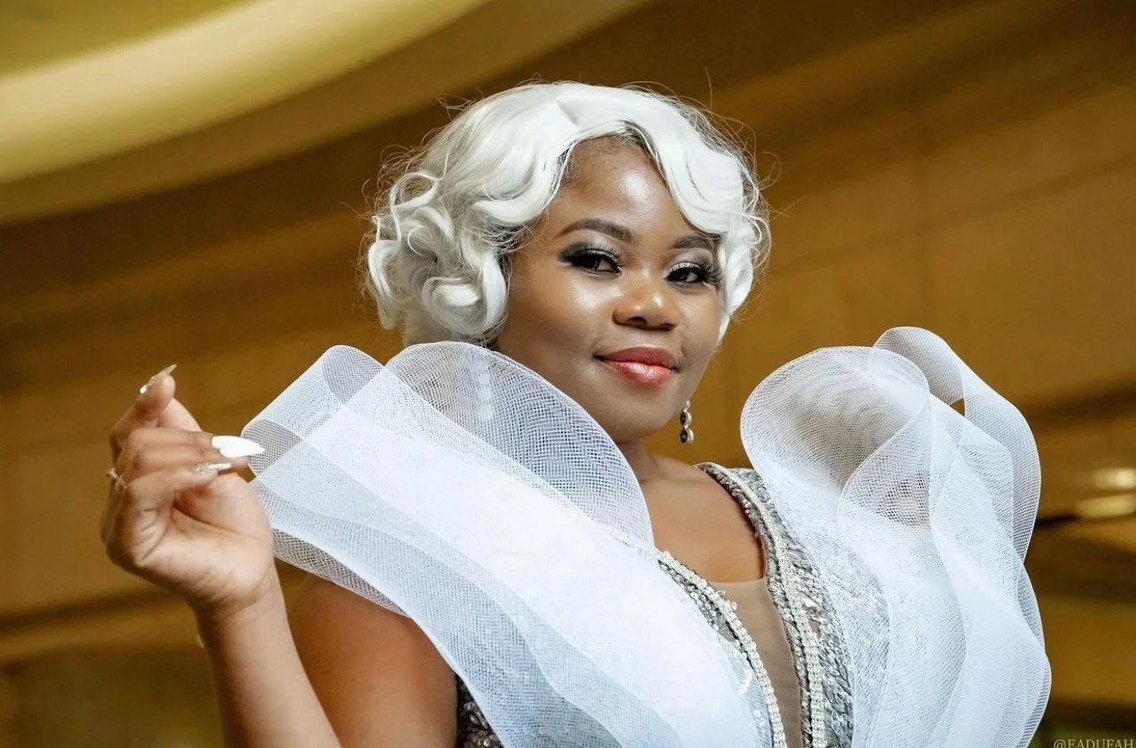 A woman with curly gray hair wearing a glamorous dress with large white ruffled shoulder features.