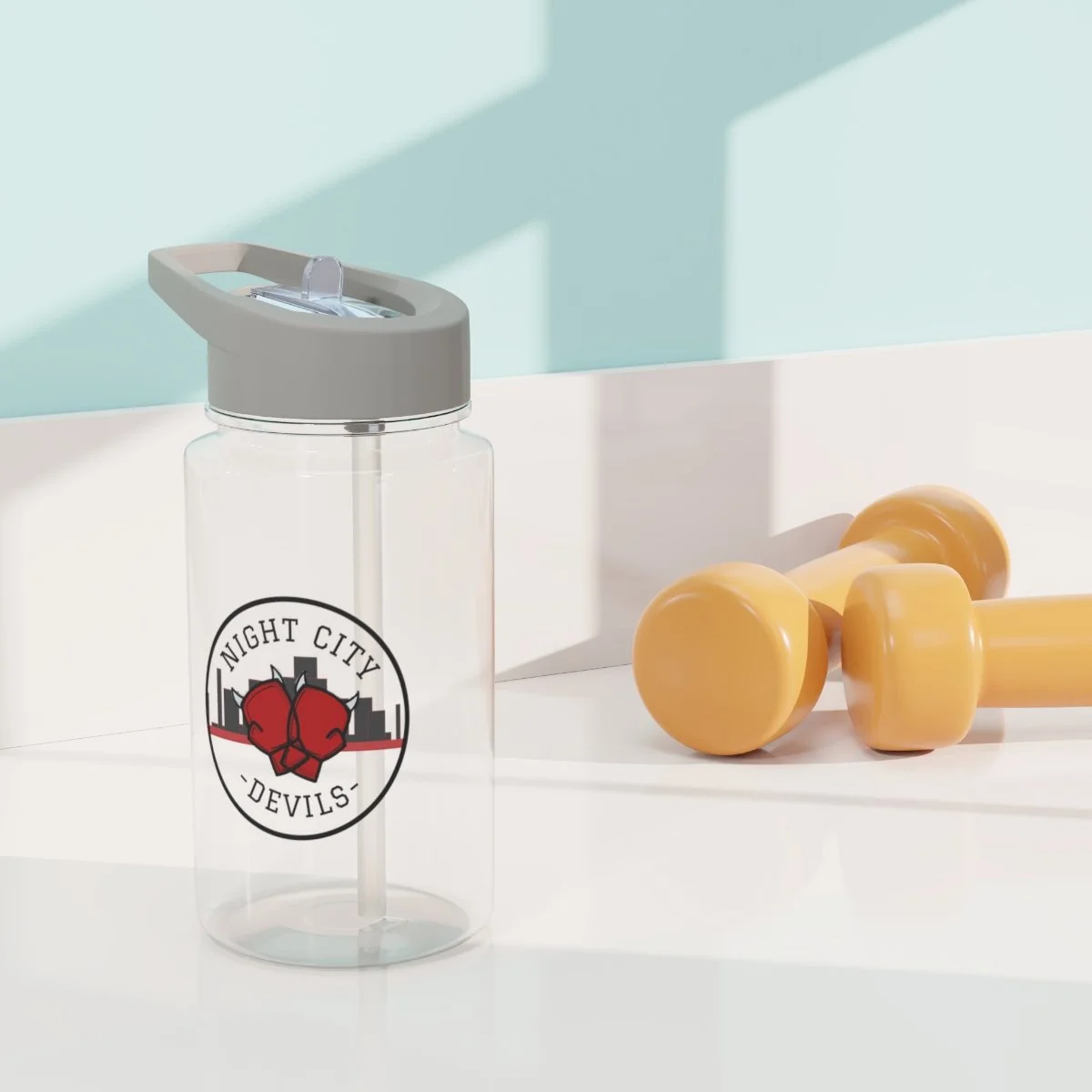 A transparent water bottle with a gray lid and a straw, featuring a logo of a boxing glove with the text "Night City Devils," beside yellow dumbbells on a white surface with a light blue wall casting shadows.