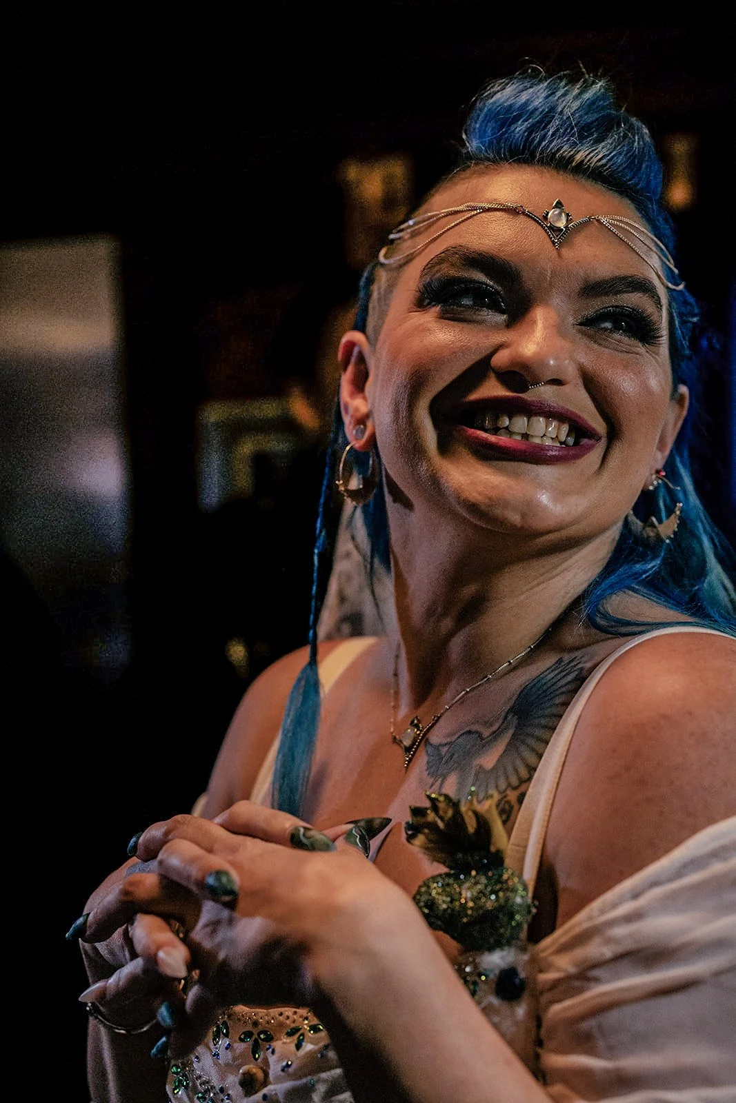A smiling woman with blue hair, tattoos, and jewelry, wearing a white top, in a dimly lit setting.