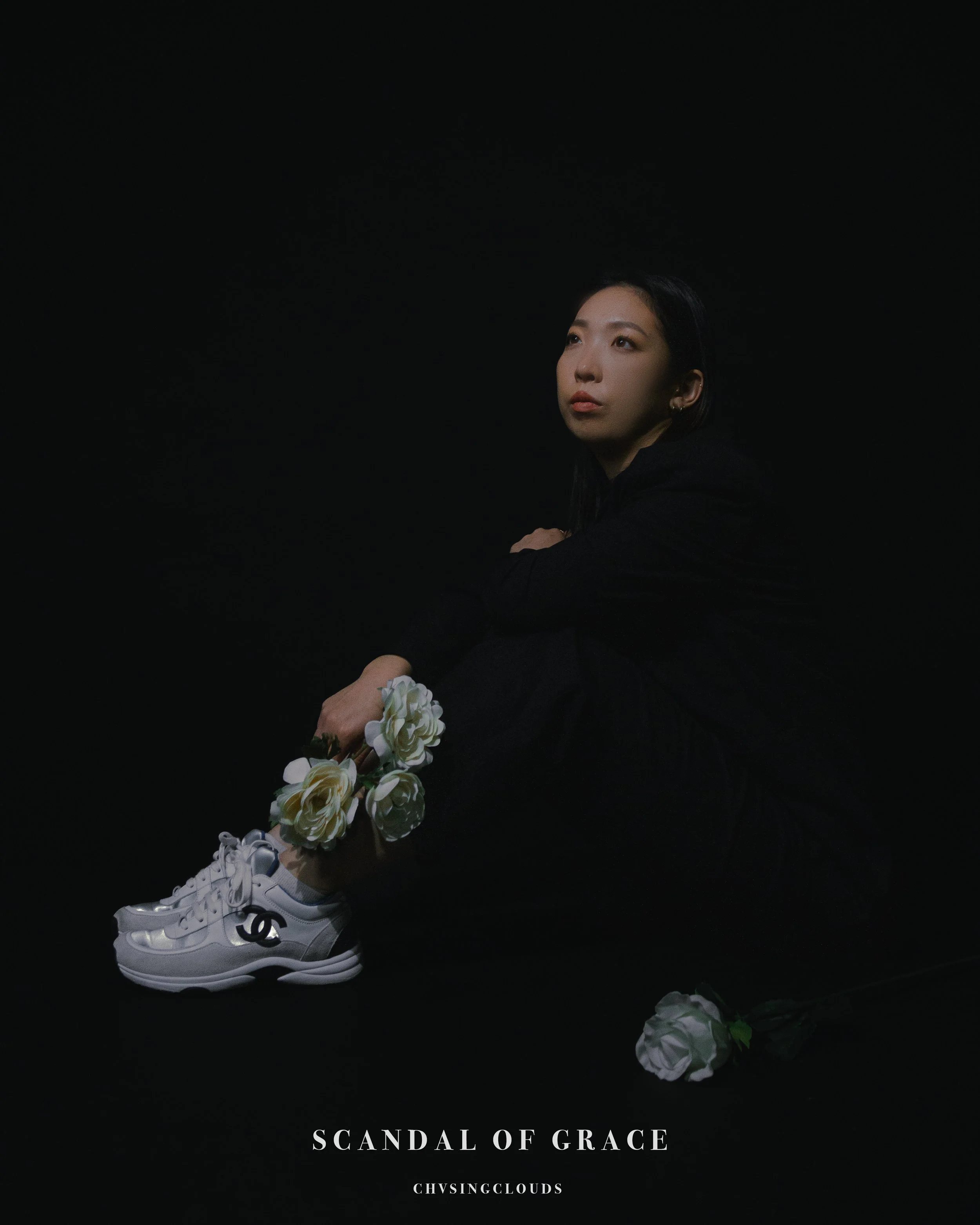 A young woman with dark hair sitting on a black backdrop, wearing a black outfit, holding white shoes with a Chanel logo, surrounded by white roses, and the words "Scandal of Grace" and "CHVISINGCLOUDS" at the bottom.