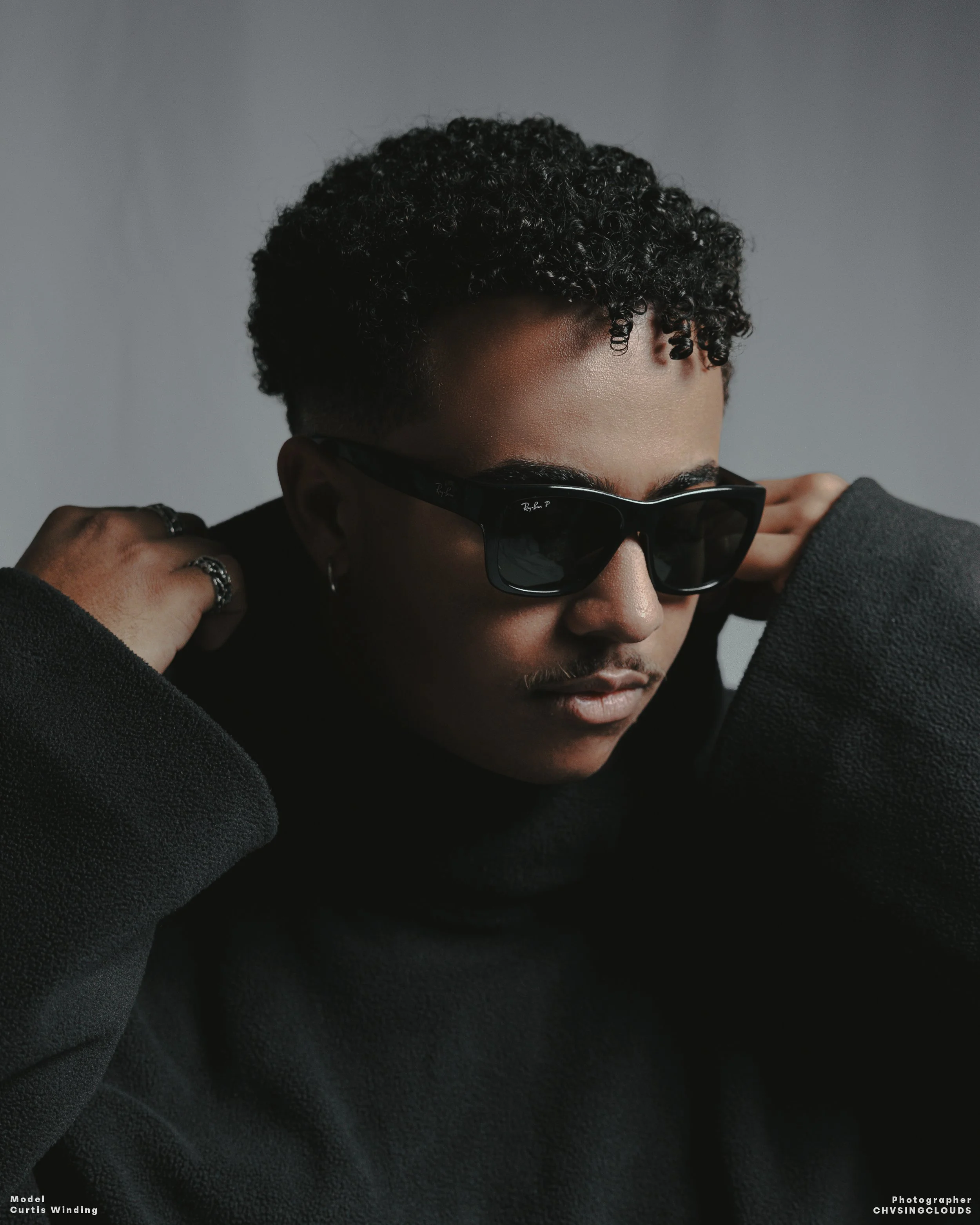 A person with short curly hair wearing sunglasses, a black sweater, and rings, posing with hands behind the neck against a plain gray background.