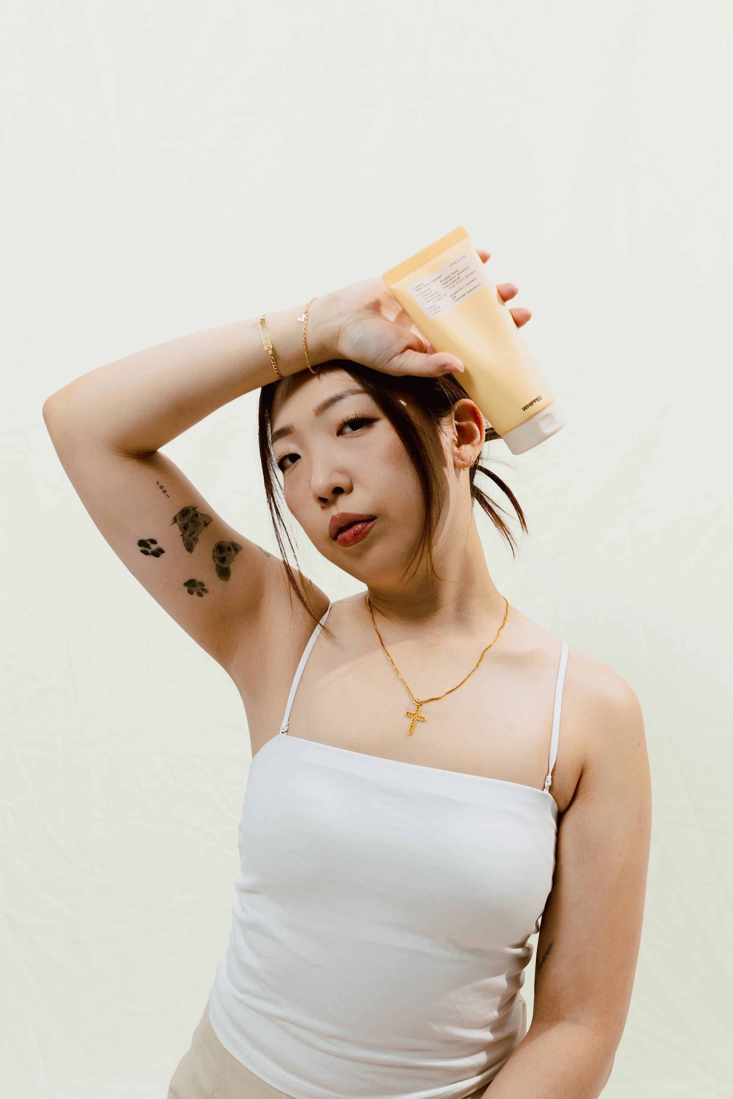 A woman with tattoos on her left arm holding a yellow bottle of lotion or sunscreen on her head, posing against a plain white wall.