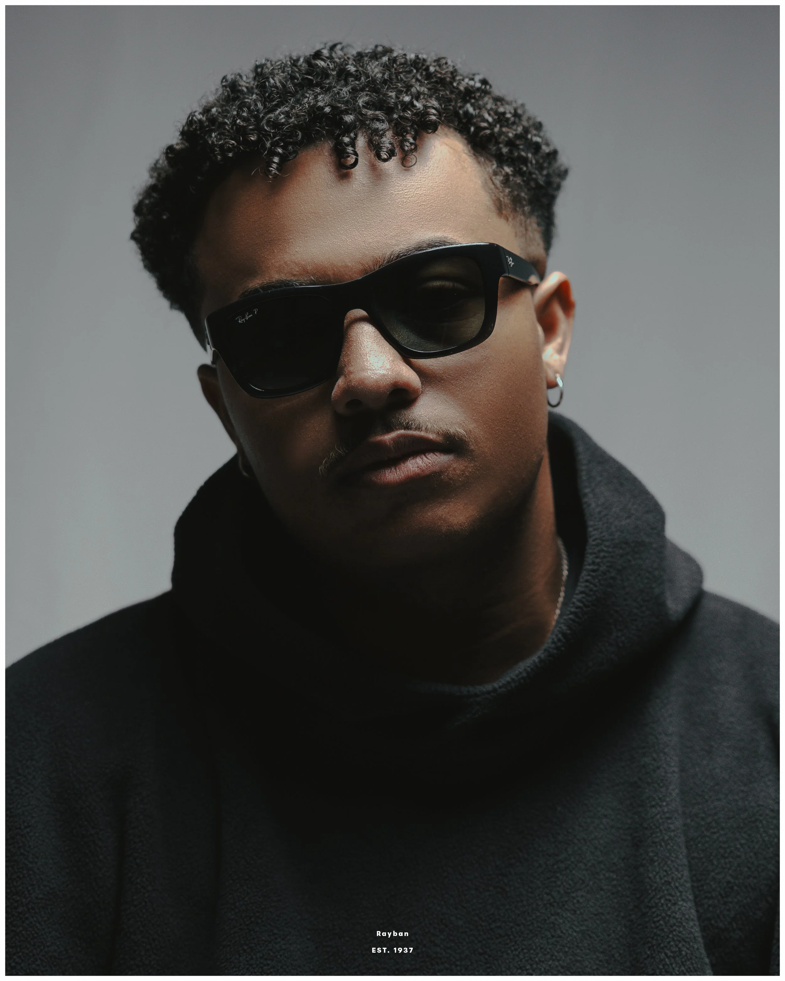 A person wearing black Ray-Ban sunglasses, a black hoodie, and small hoop earrings, with a neutral expression against a plain gray background.